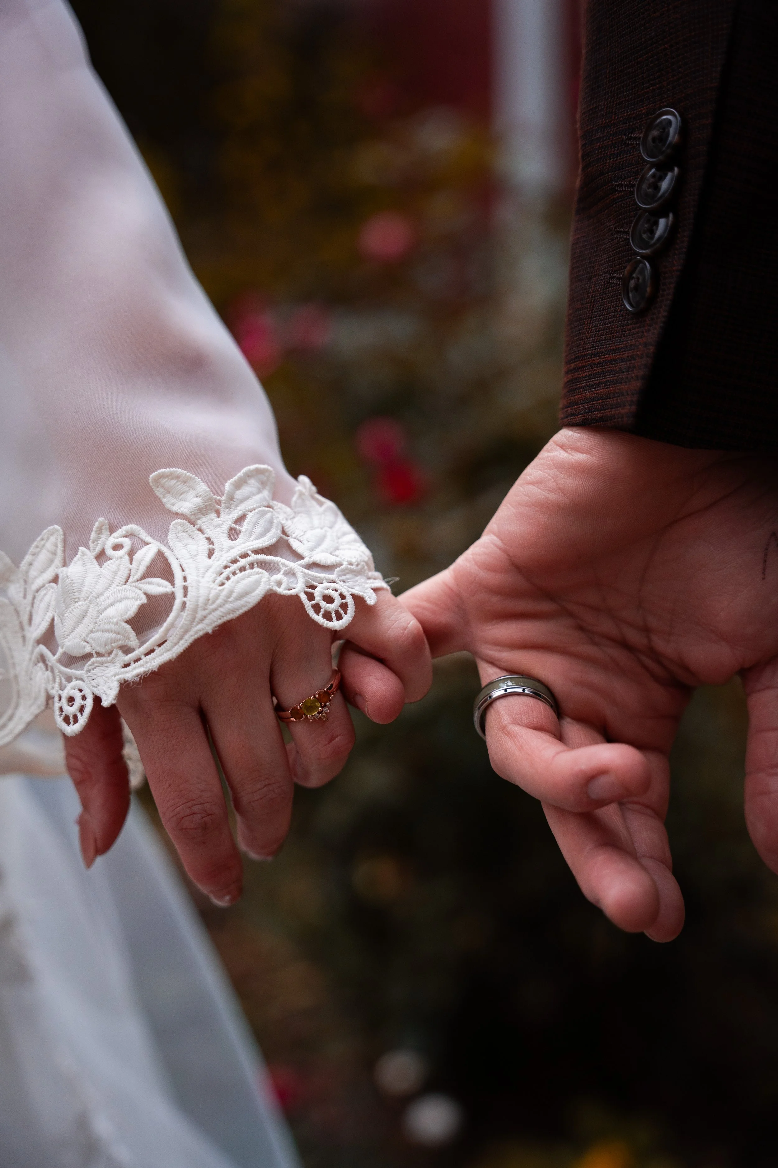 Close-up of a bride and groom holding hands, with the bride's hand on top, showing wedding rings, and the groom's hand underneath. The bride's clothing features lace embroidery, and the background has a blurred outdoor setting.