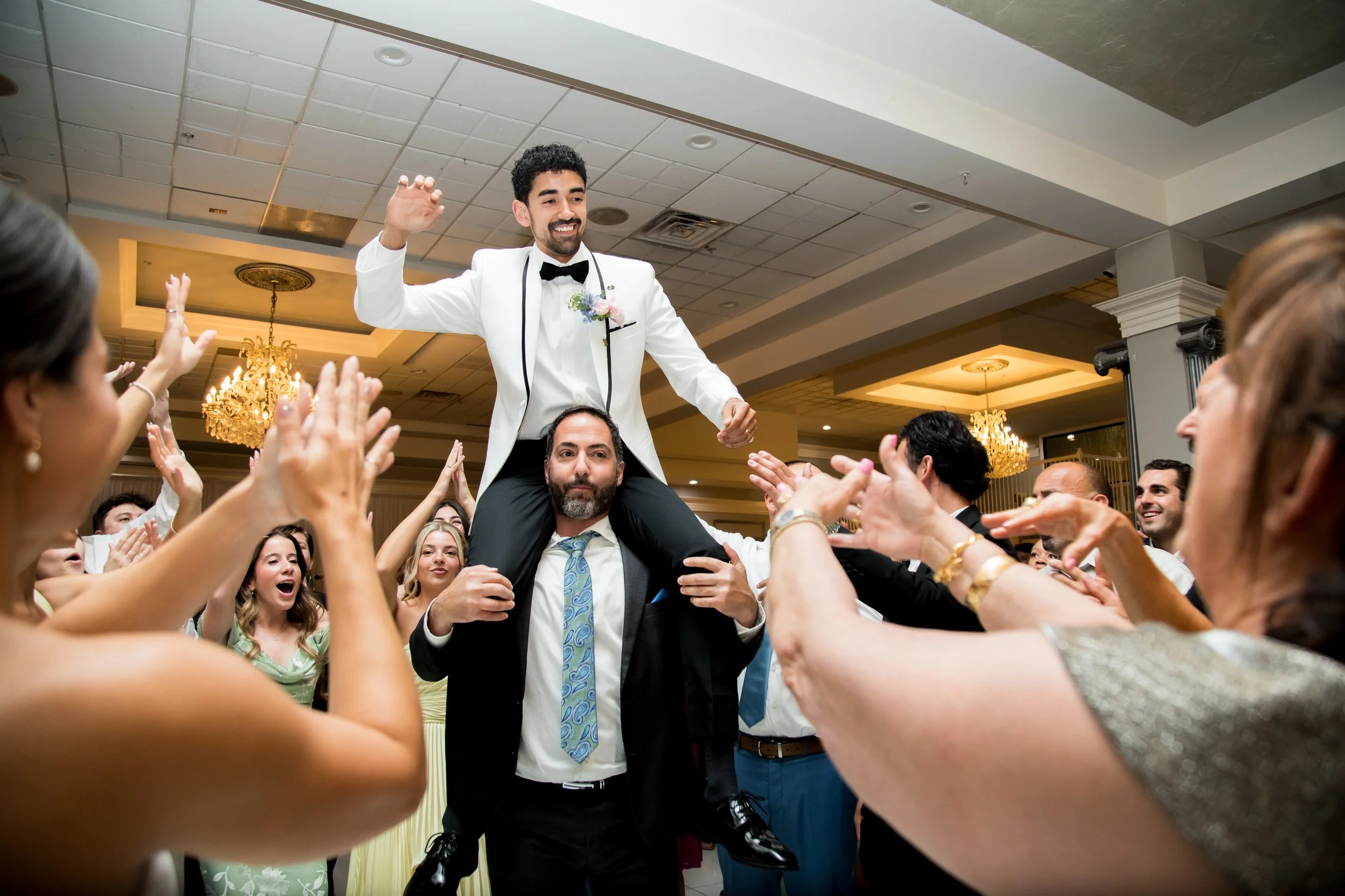 A groom in a white tuxedo with a bow tie and boutonniere stands on the shoulders of a man in a suit during a wedding celebration. He is surrounded by applauding guests, many with their hands raised. The setting is an elegant indoor banquet hall with 