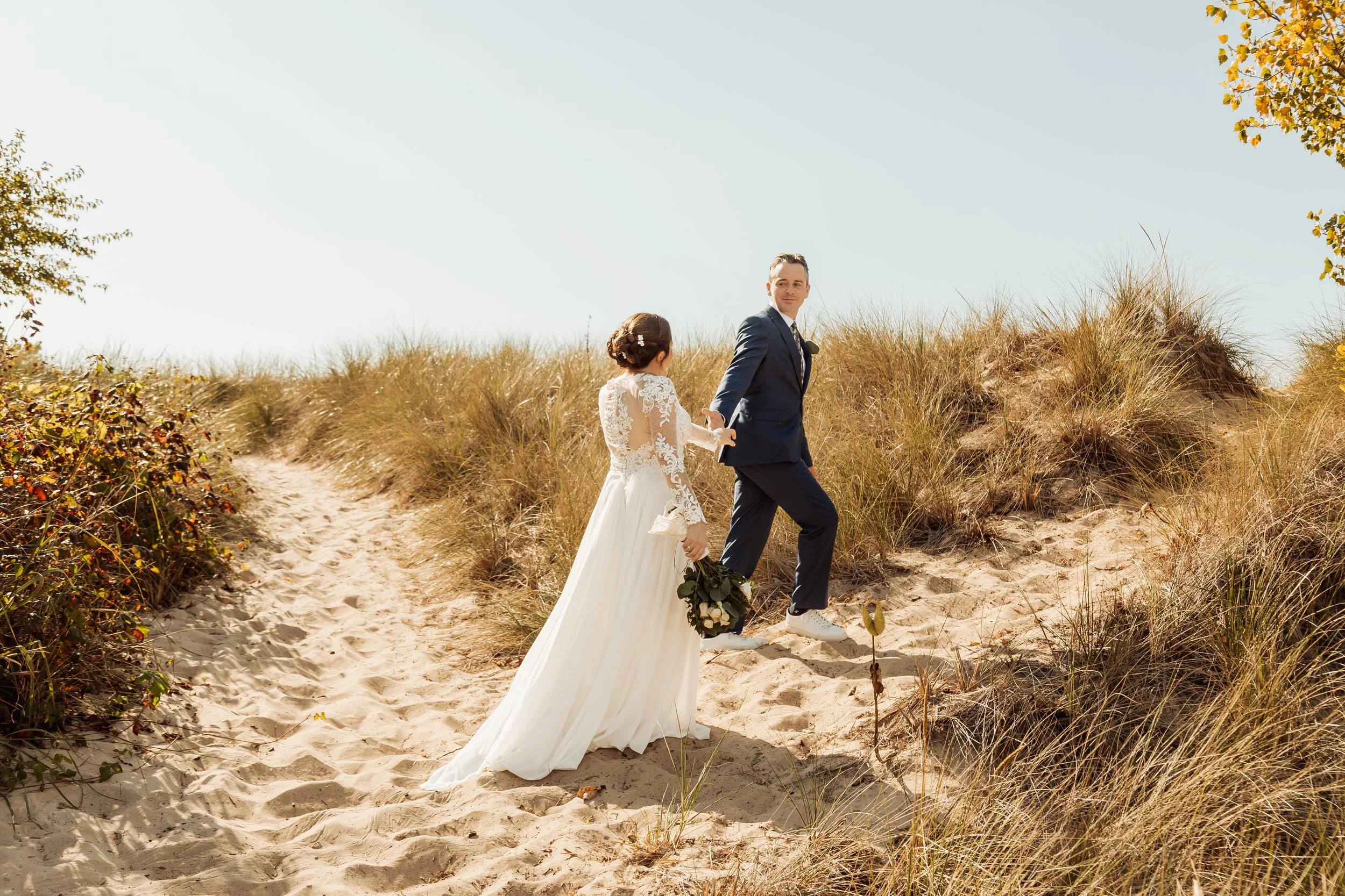 A bride and groom walking hand in hand on sandy dunes with desert vegetation and a clear sky.