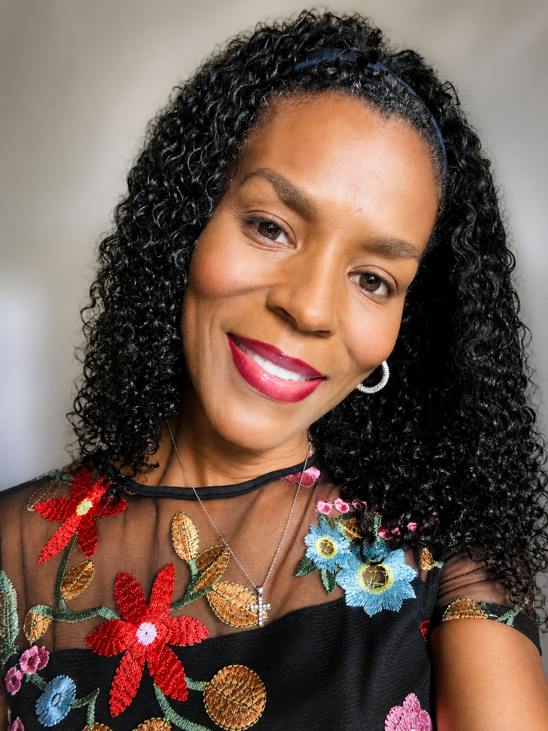 A woman with curly black hair wearing a floral embroidered sheer top, a necklace with a cross pendant, and hoop earrings, smiling at the camera.