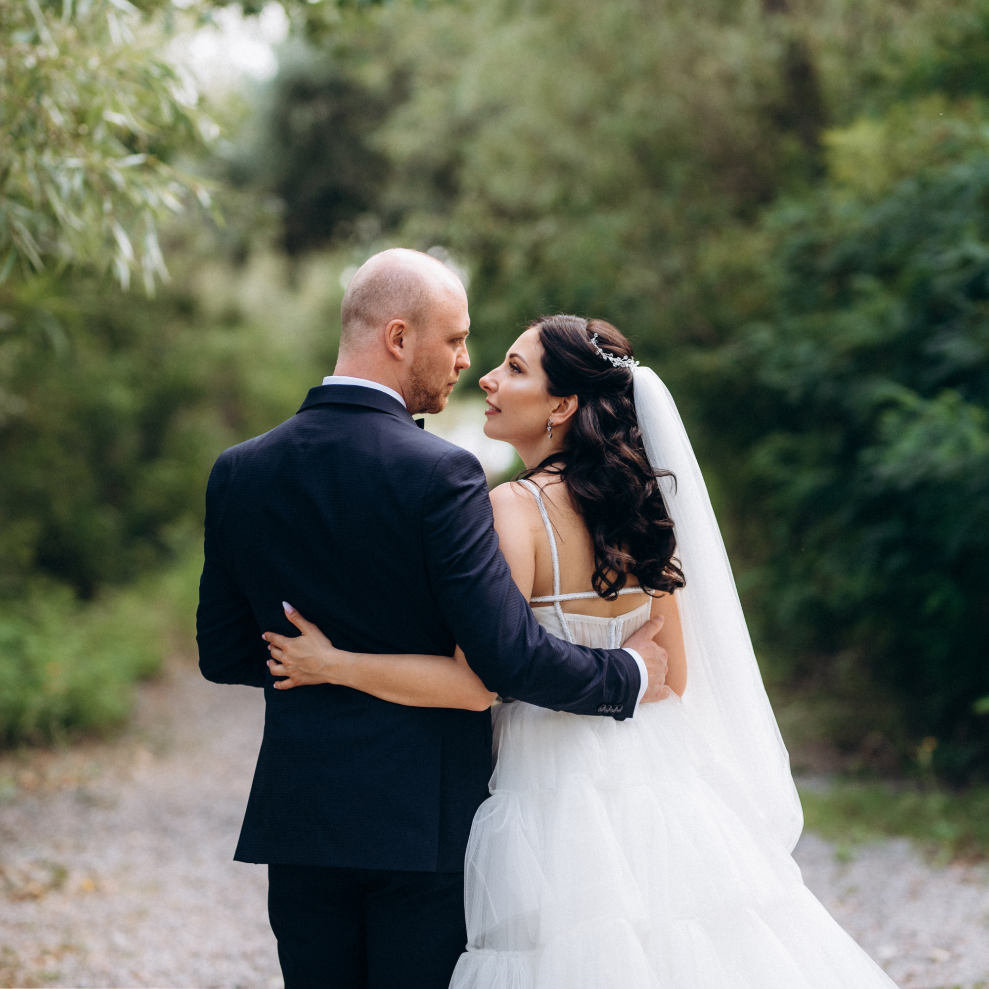 A bride and groom standing close together outdoors surrounded by trees, looking into each other's eyes during their wedding.