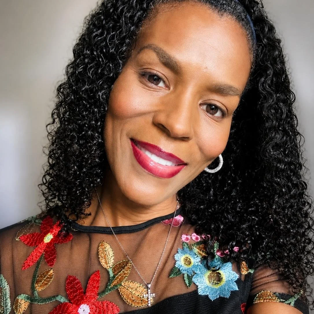 Close-up of a woman with dark curly hair, wearing red lipstick, a floral embroidered sheer top, and jewelry, smiling at the camera.