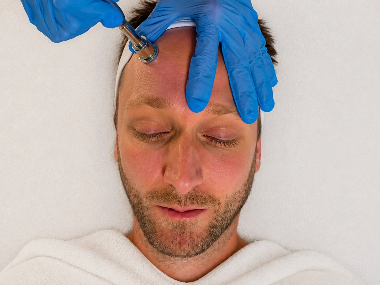 Mann liegt entspannt mit geschlossenen Augen im Kosmetikstudion Hautidee in Schwülper, während eine Person mit medizinischen Schutzhandschuhen eine Microdermabrasion an der Stirn des Mannes durchführt.