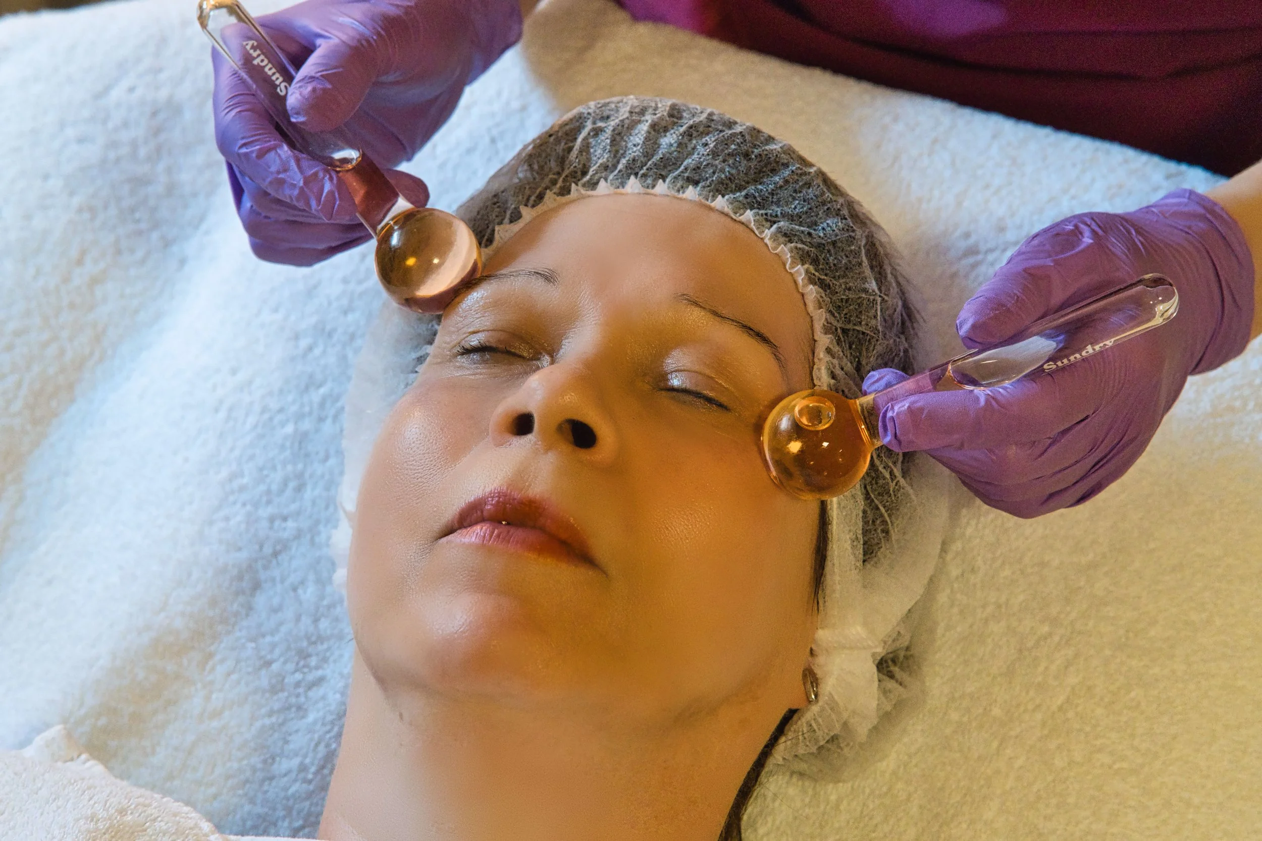 Person erhält eine Gesichtsbehandlung Das Kosmetikstudio Hautidee in Schwülper mit einem kosmetischen Werkzeug, trägt eine Haarhaube, liegt auf einer Behandlungsliege, während eine Kosmetikerin mit lilafarbenen Handschuhen arbeitet.