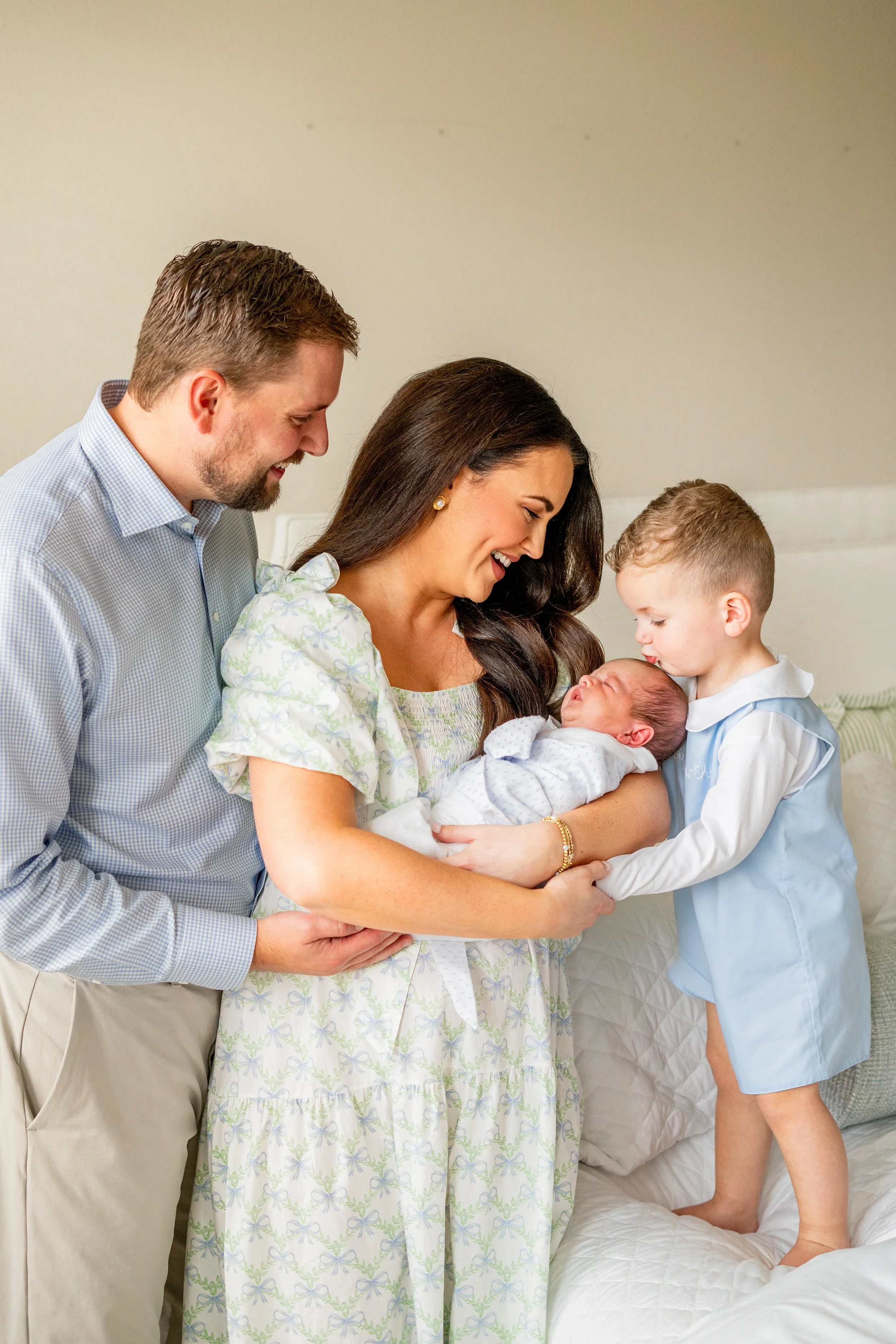 A family in a hospital room celebrating a newborn baby. The mother holds the baby, while a young boy kisses the newborn on the head, and a man looks on smiling.