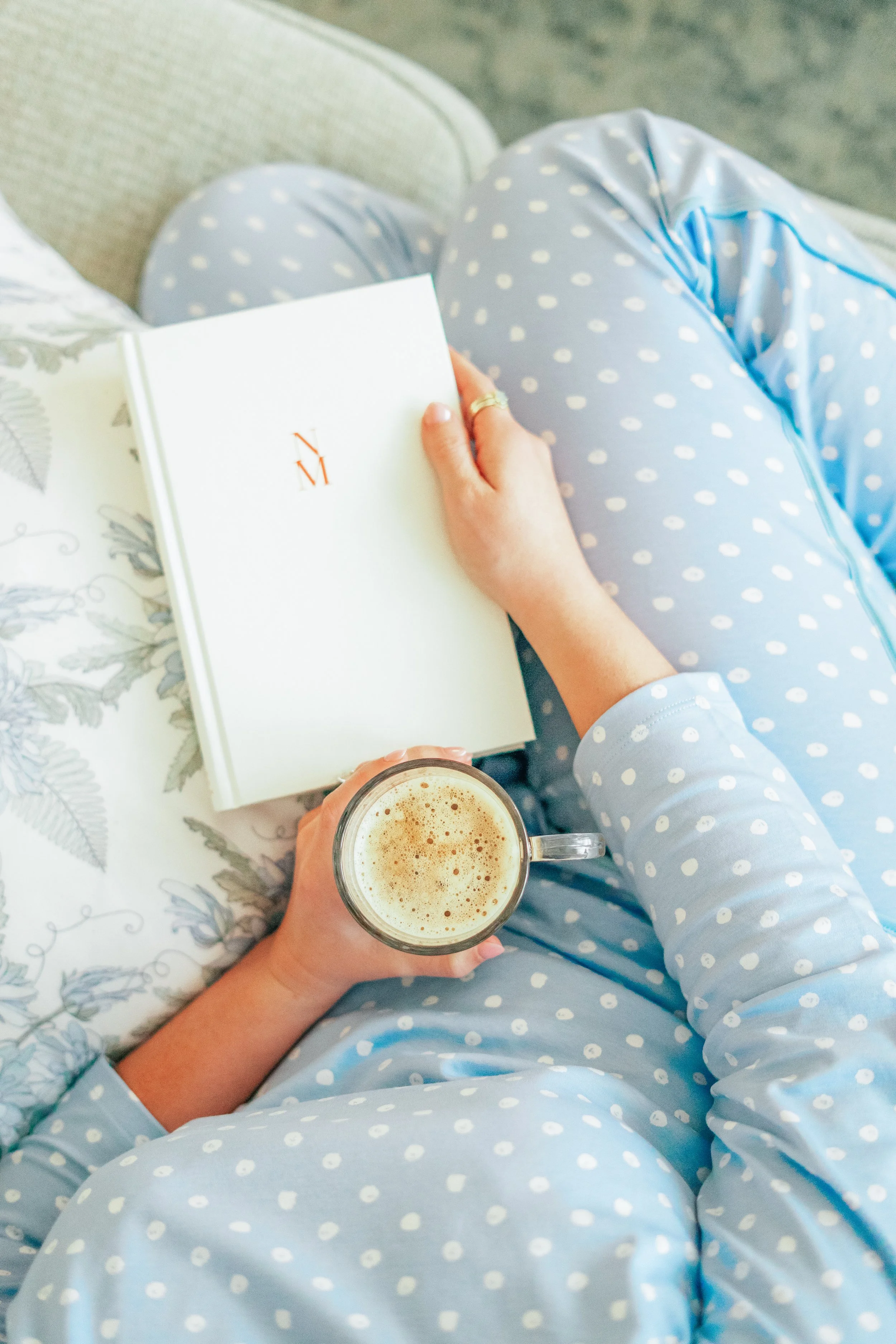 Person wearing light blue polka dot pajamas sitting on a couch, holding a book and a glass of coffee, with a floral cushion nearby.