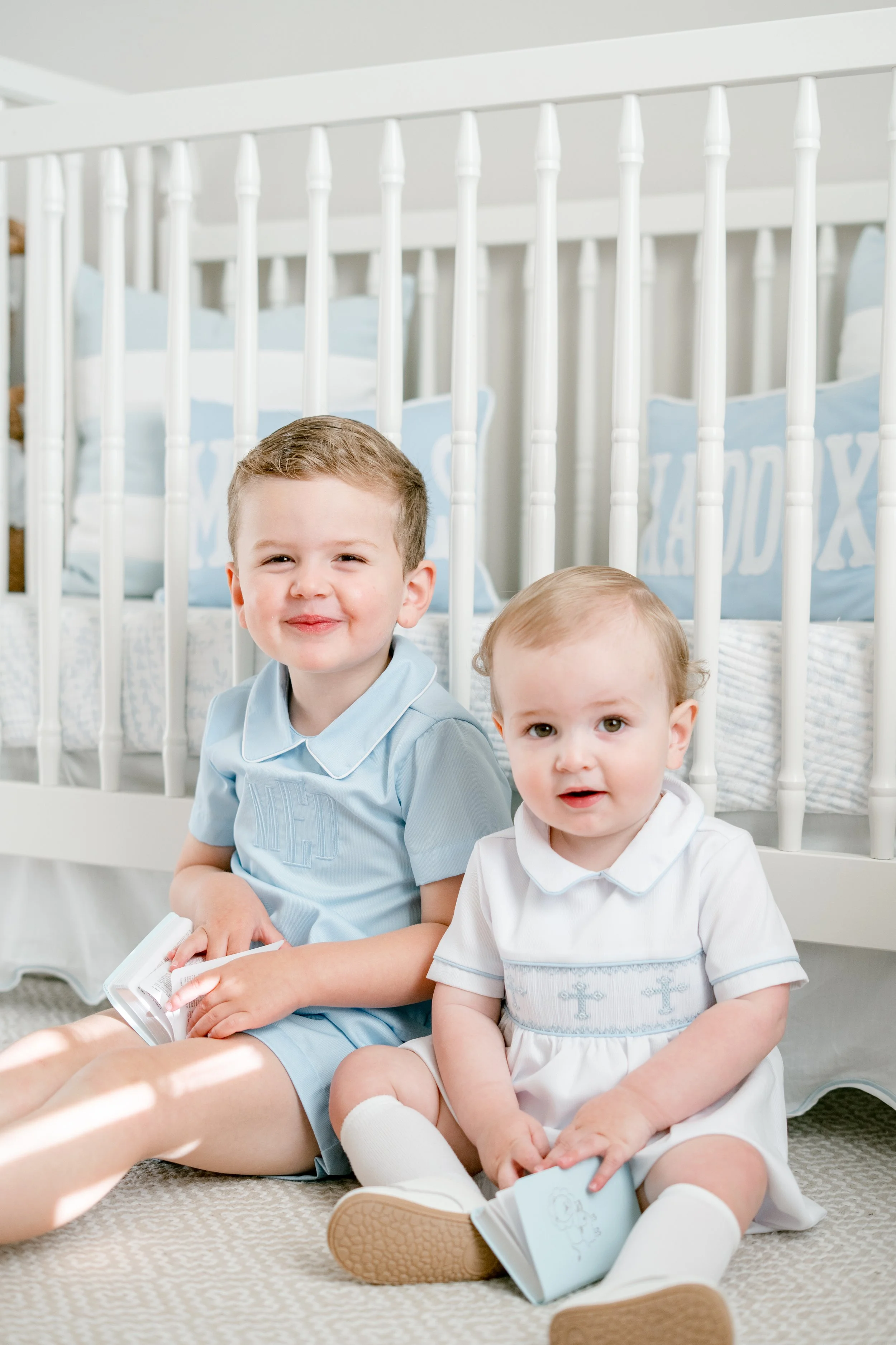Two young boys sitting on a carpeted floor in front of a white bed with a white safety rail, smiling and holding books in a bright bedroom.
