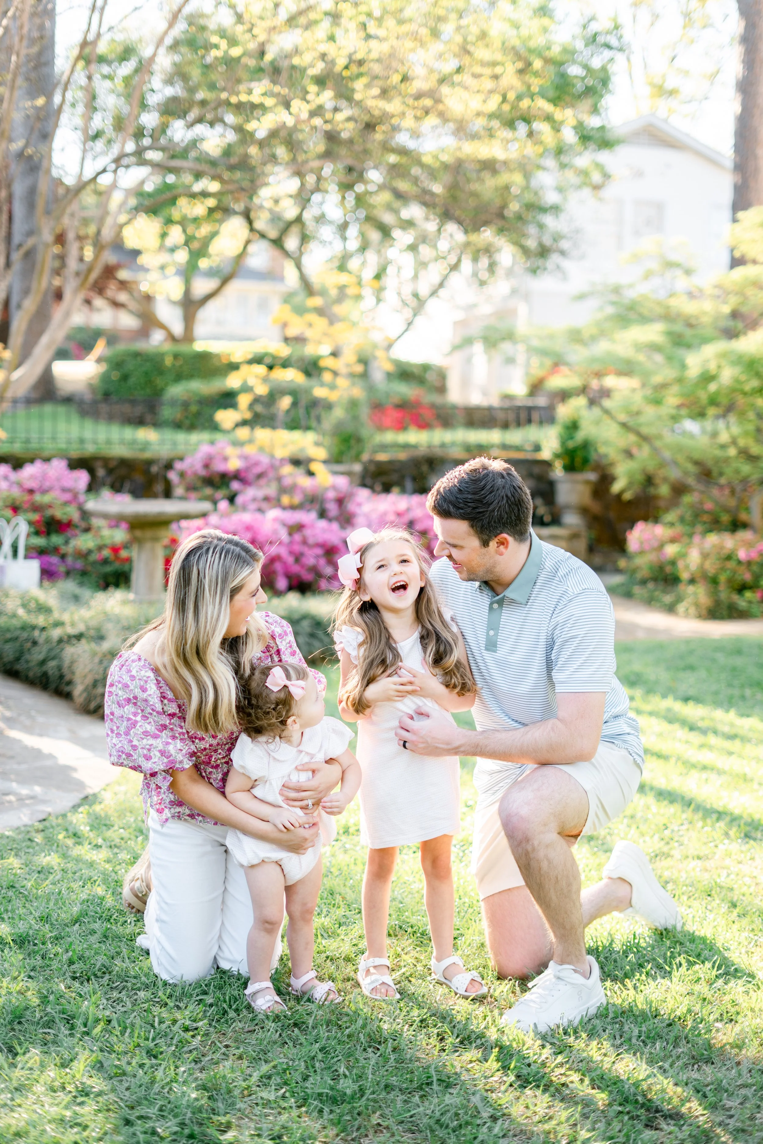 A family of four, including two young girls and their parents, is outdoors in a park during daylight. They are smiling and laughing, with the father kneeling and the mother holding a girl. The background shows greenery, pink flowers, and a sunny sky.