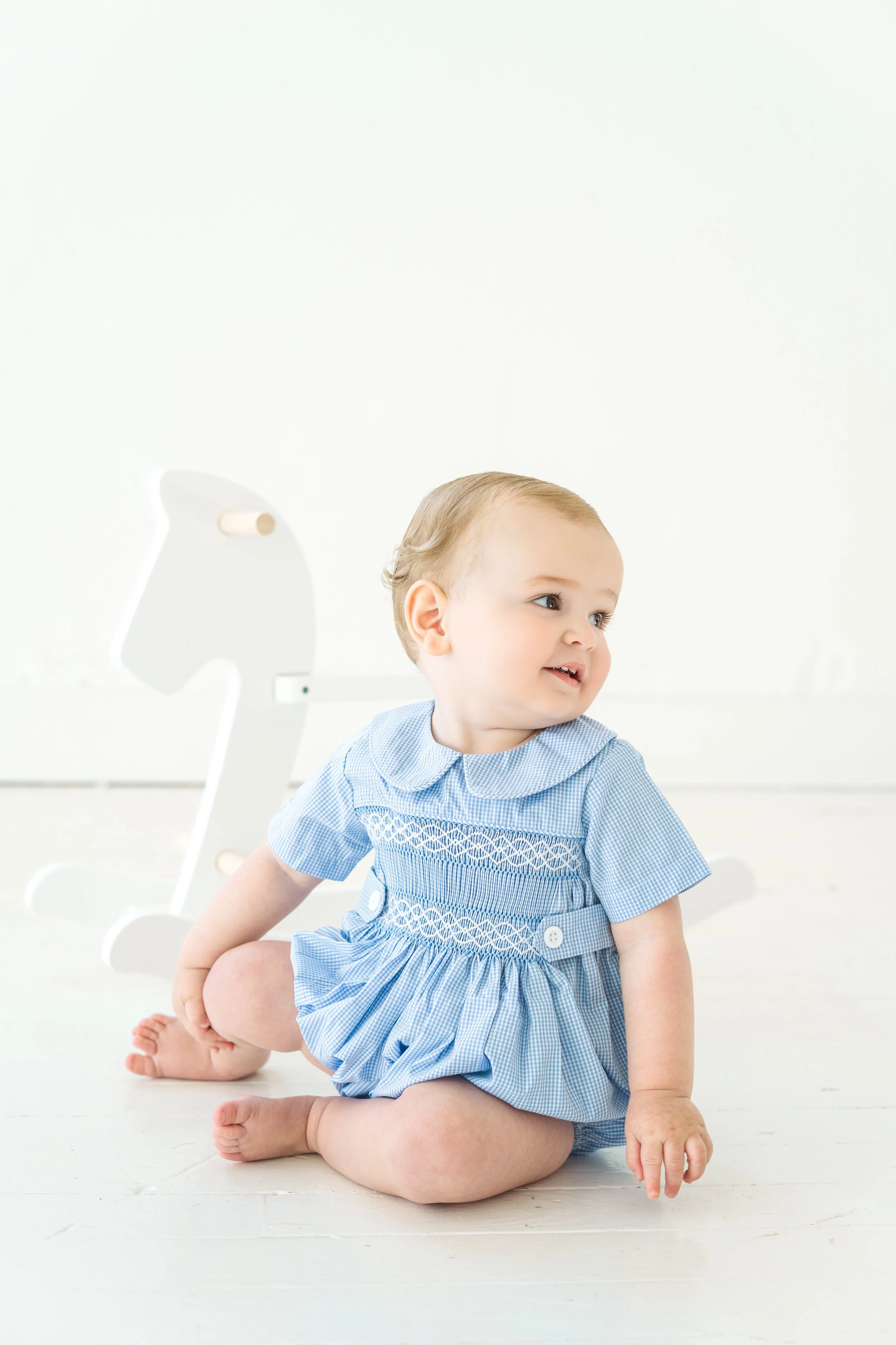 A young child with blonde hair, blue eyes, and wearing a blue and white checkered dress, sitting on a light-colored floor with a white background and a lamp in the background.