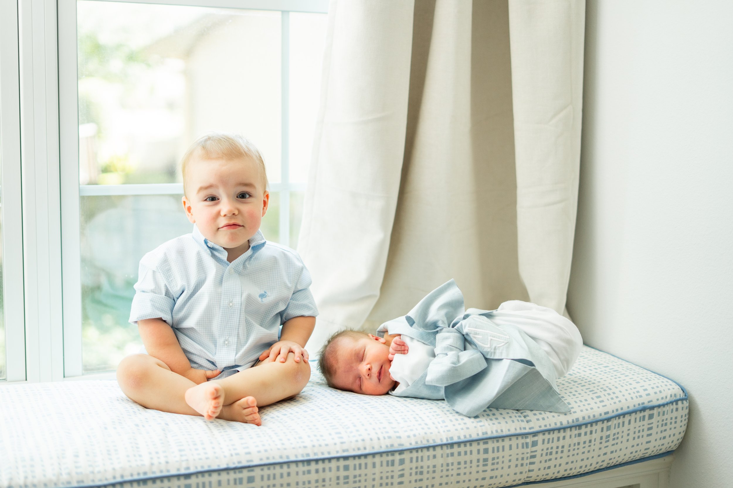 A young boy with light skin, blonde hair, and blue eyes sitting on a bed next to a sleeping newborn baby, near a large window with curtains, in a bright room.