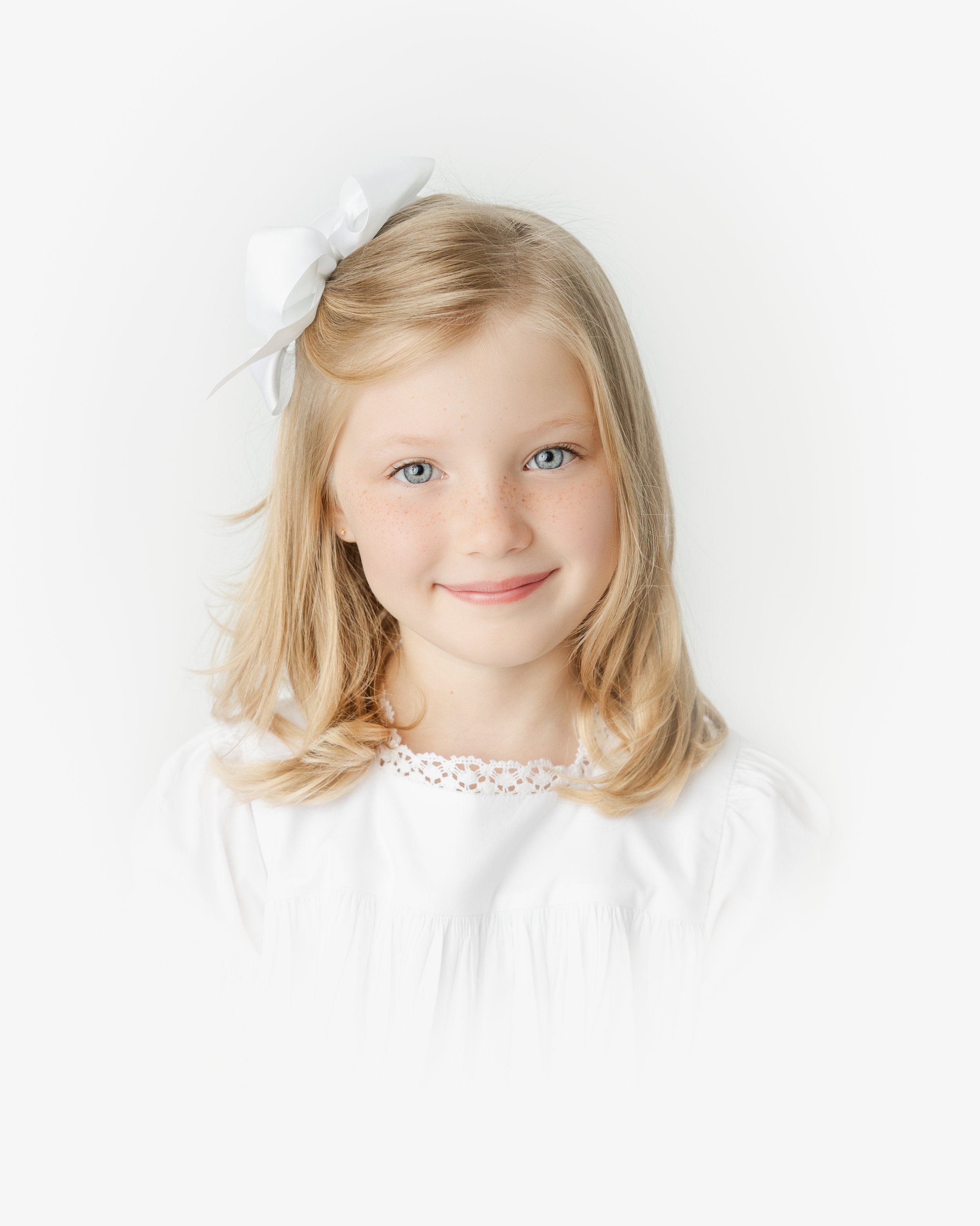 Close-up portrait of a young girl with blonde hair, fair skin, blue eyes, and a gentle smile, wearing a white dress with lace detail and a large white bow in her hair, against a plain white background.