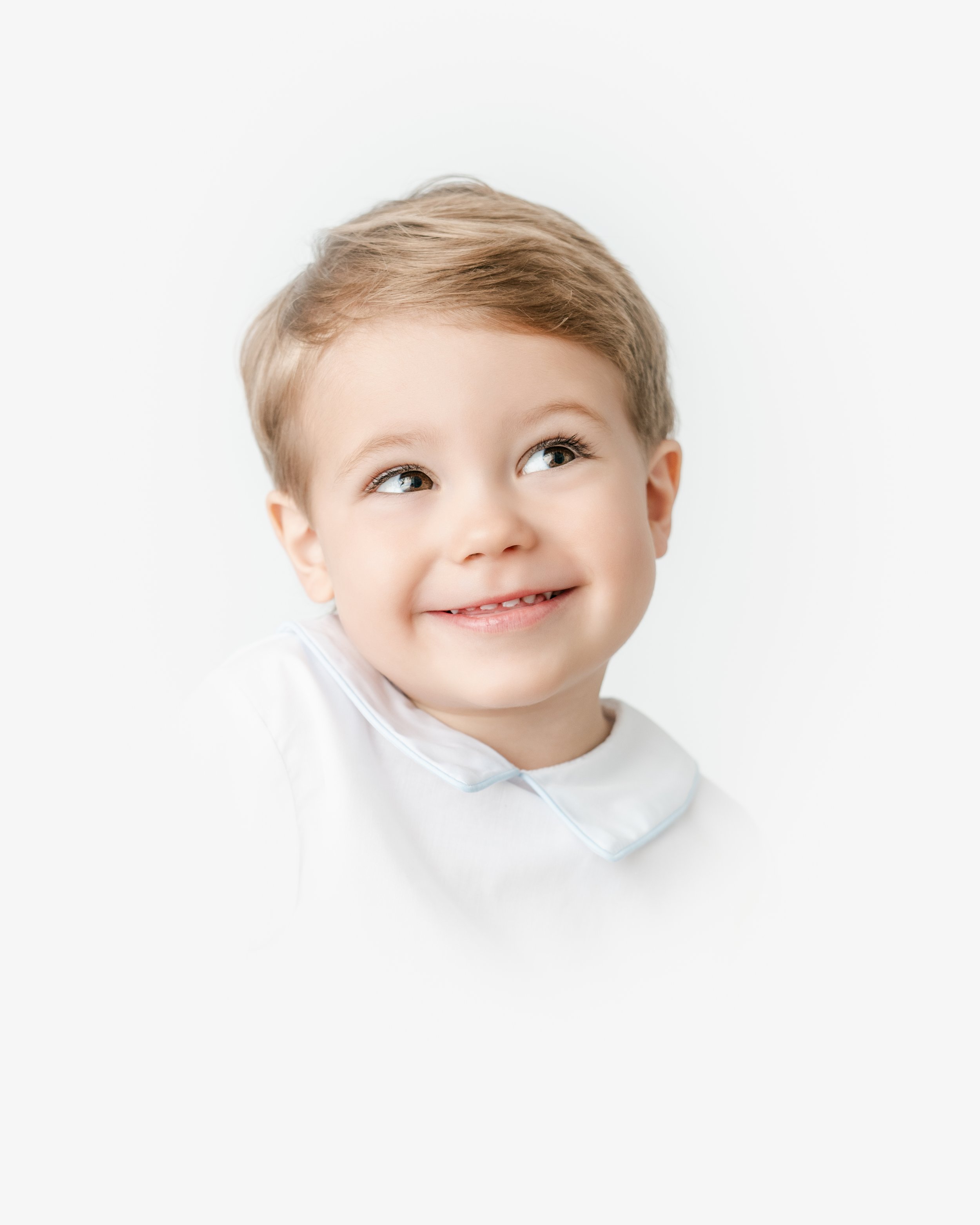 Close-up of a young boy with short blonde hair and brown eyes smiling and looking upwards against a white background.