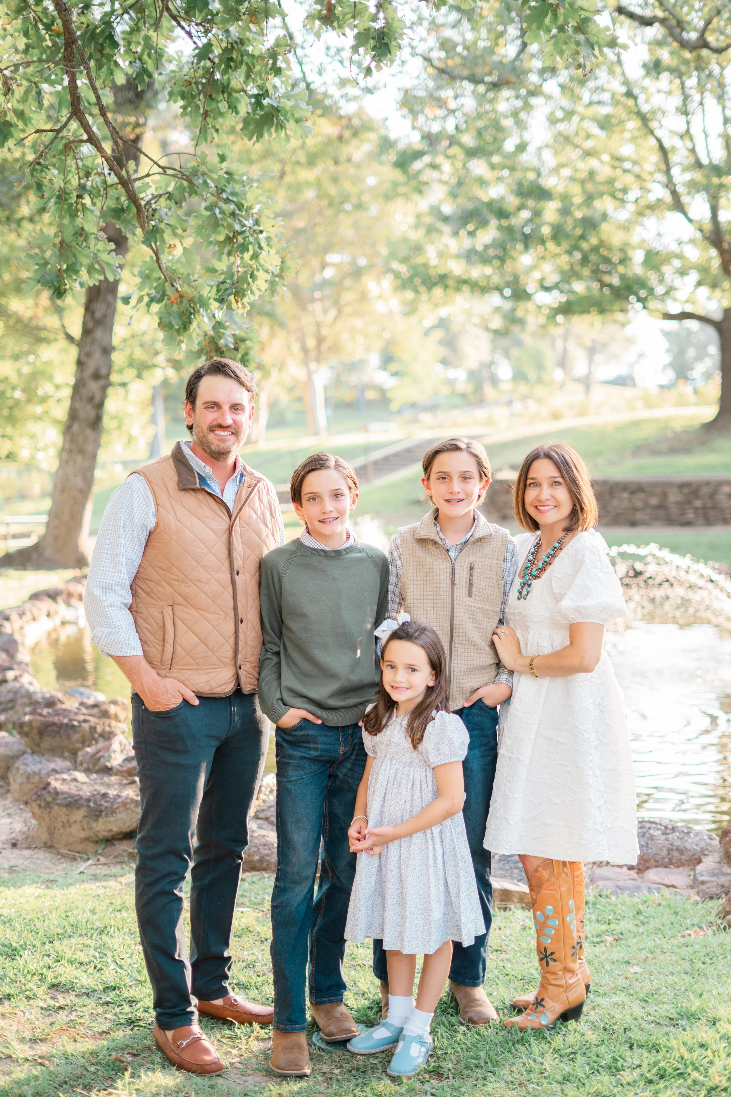 A family of six standing outdoors near a pond with trees and sunlight. The group includes two adults and four children, all smiling and dressed casually.