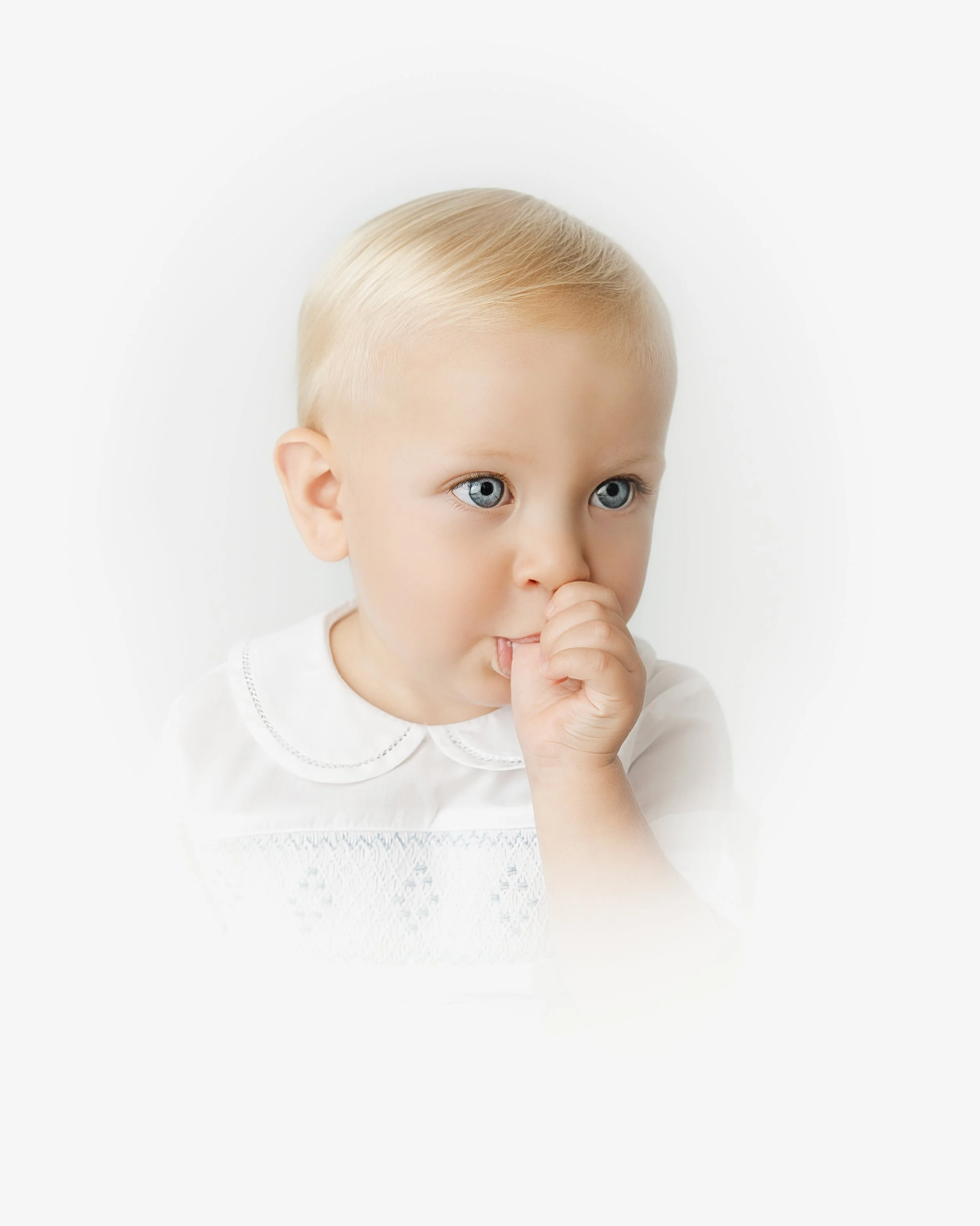 Close-up of a young child with blonde hair and blue eyes, wearing a white shirt, holding their finger to their lips in a thoughtful pose.