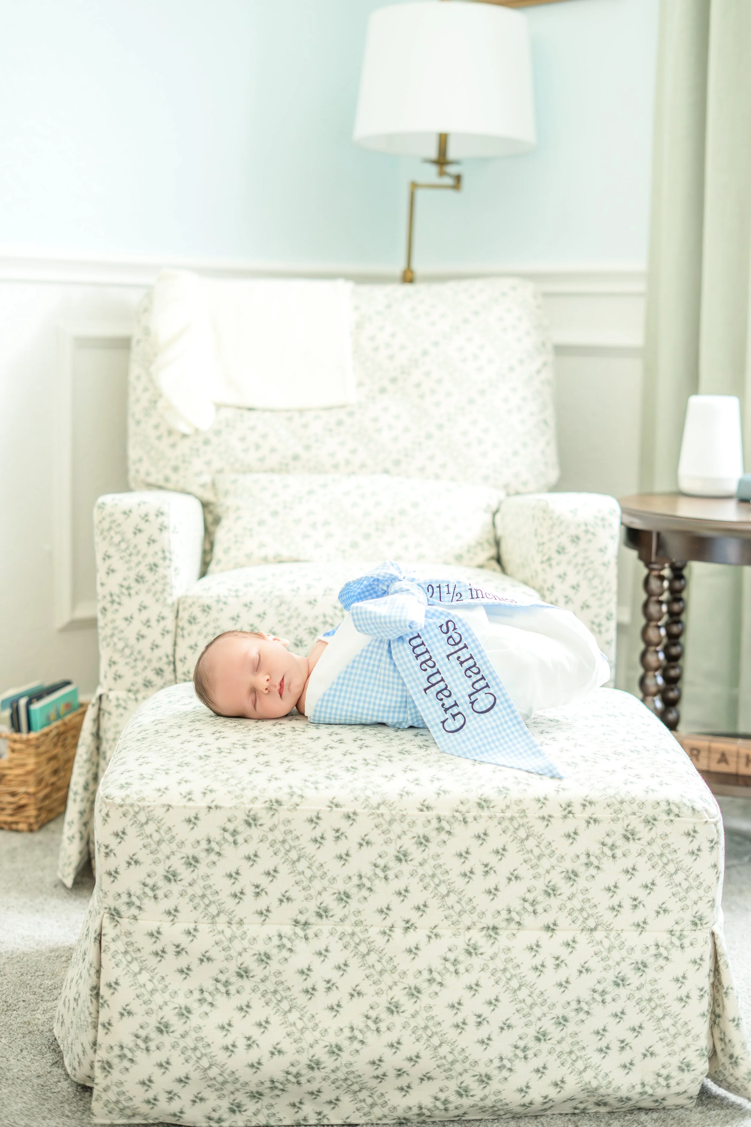 A sleeping baby dressed in a blue and white checkered outfit wrapped in a white blanket with blue trim, lying on a floral-patterned bedspread in a cozy bedroom with a beige armchair, side table with a white lamp, and a small bookshelf.
