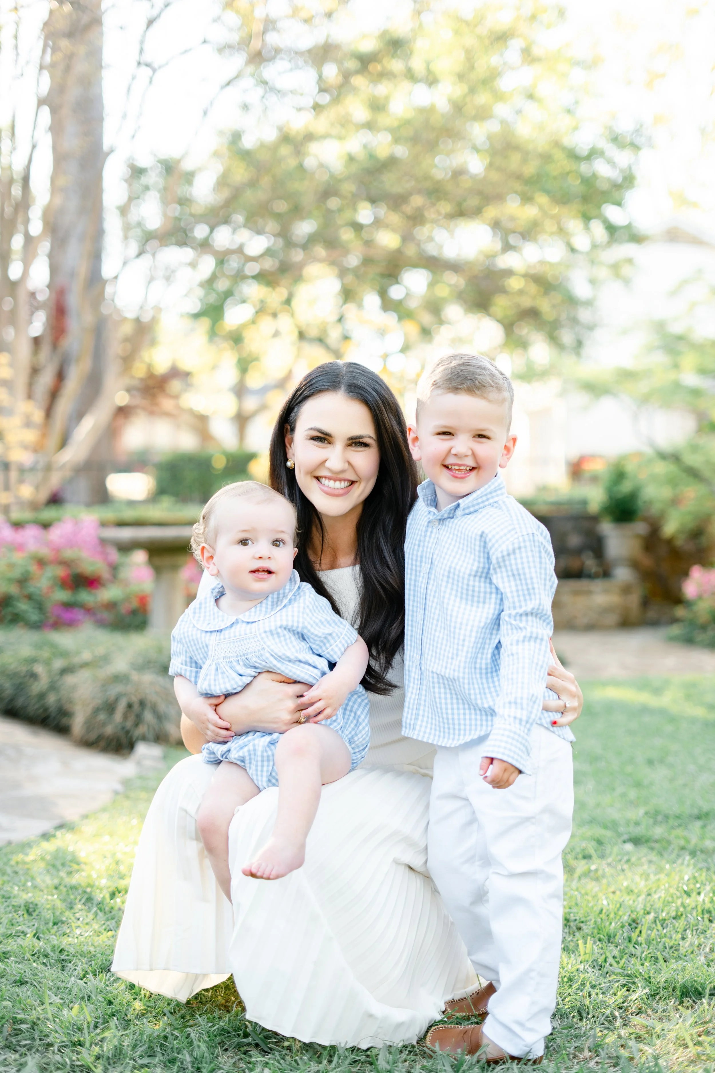 A smiling woman with long dark hair sitting outdoors on grass, holding a young girl with blonde hair, and a young boy with short light brown hair standing beside her, surrounded by greenery and flowers on a sunny day.