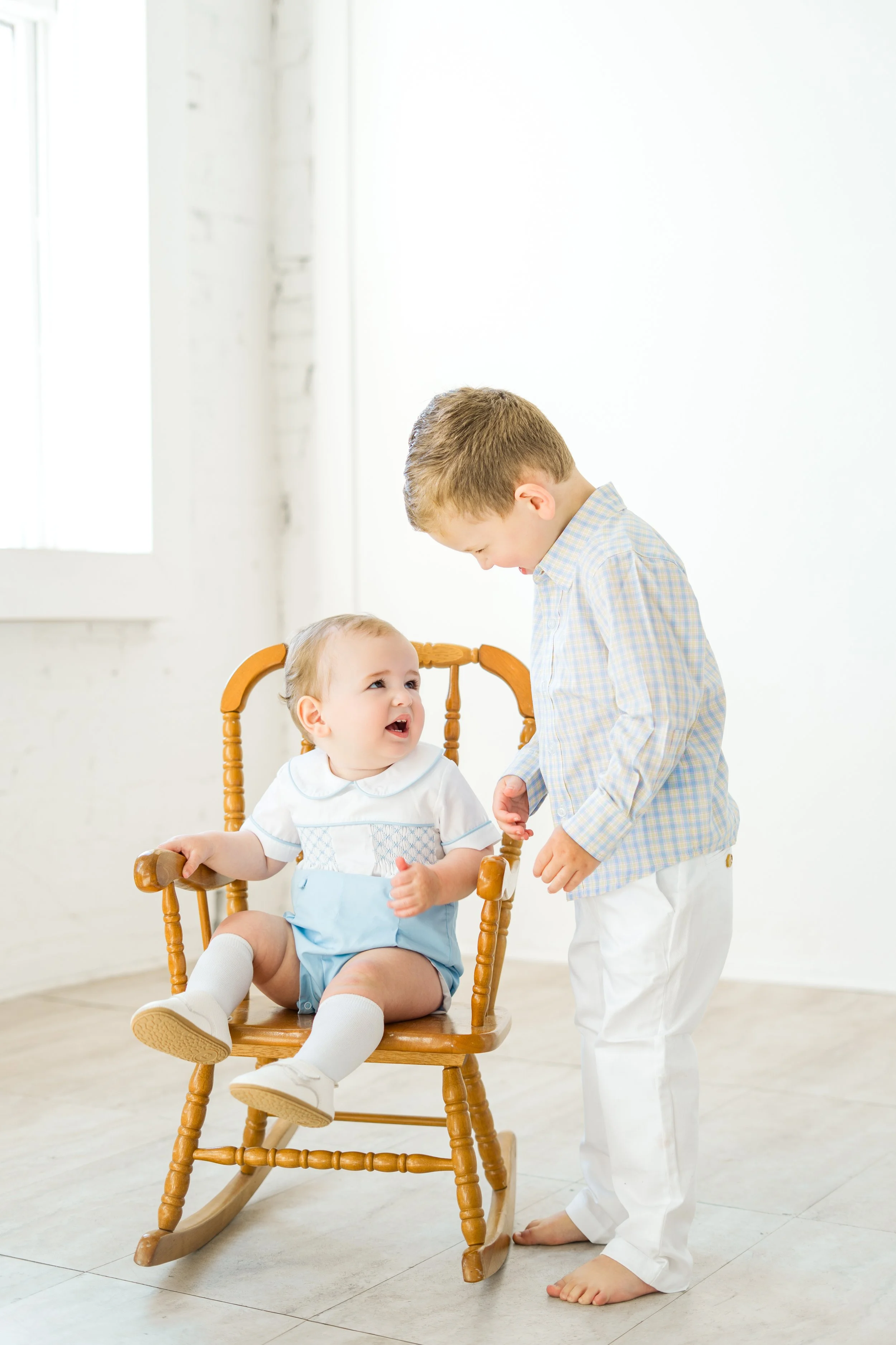 Two young boys, one sitting in a wooden rocking chair and the other standing in front of him. The boy sitting in the chair is smiling and wearing a white shirt with a pattern and light blue shorts. The standing boy is leaning forward, smiling, and we