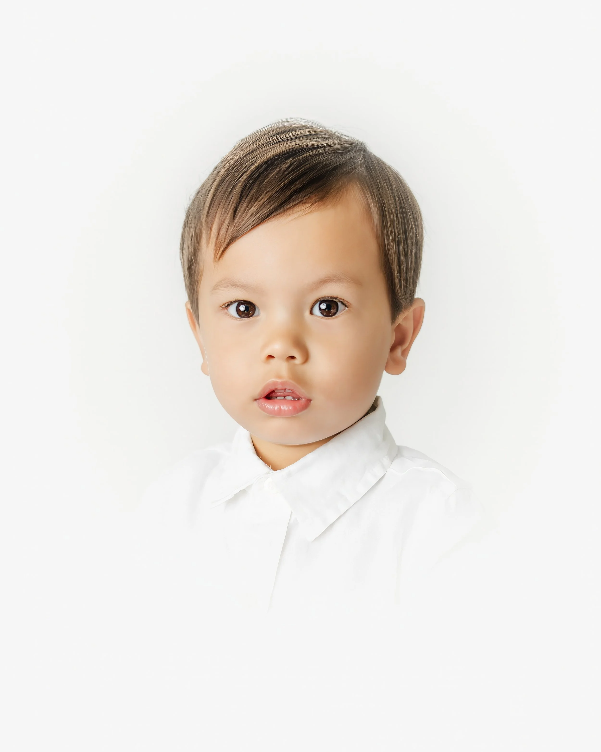 Close-up portrait of a young child with short brown hair, wearing a white shirt, against a plain white background.