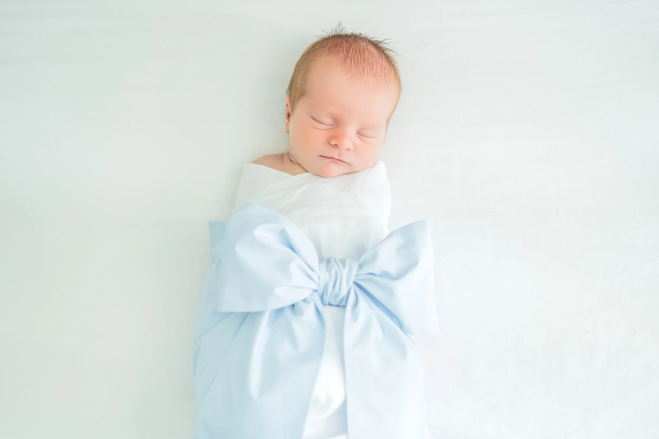 A sleeping newborn baby with red hair, wrapped in a white blanket with a large light blue bow, lying on a white surface.
