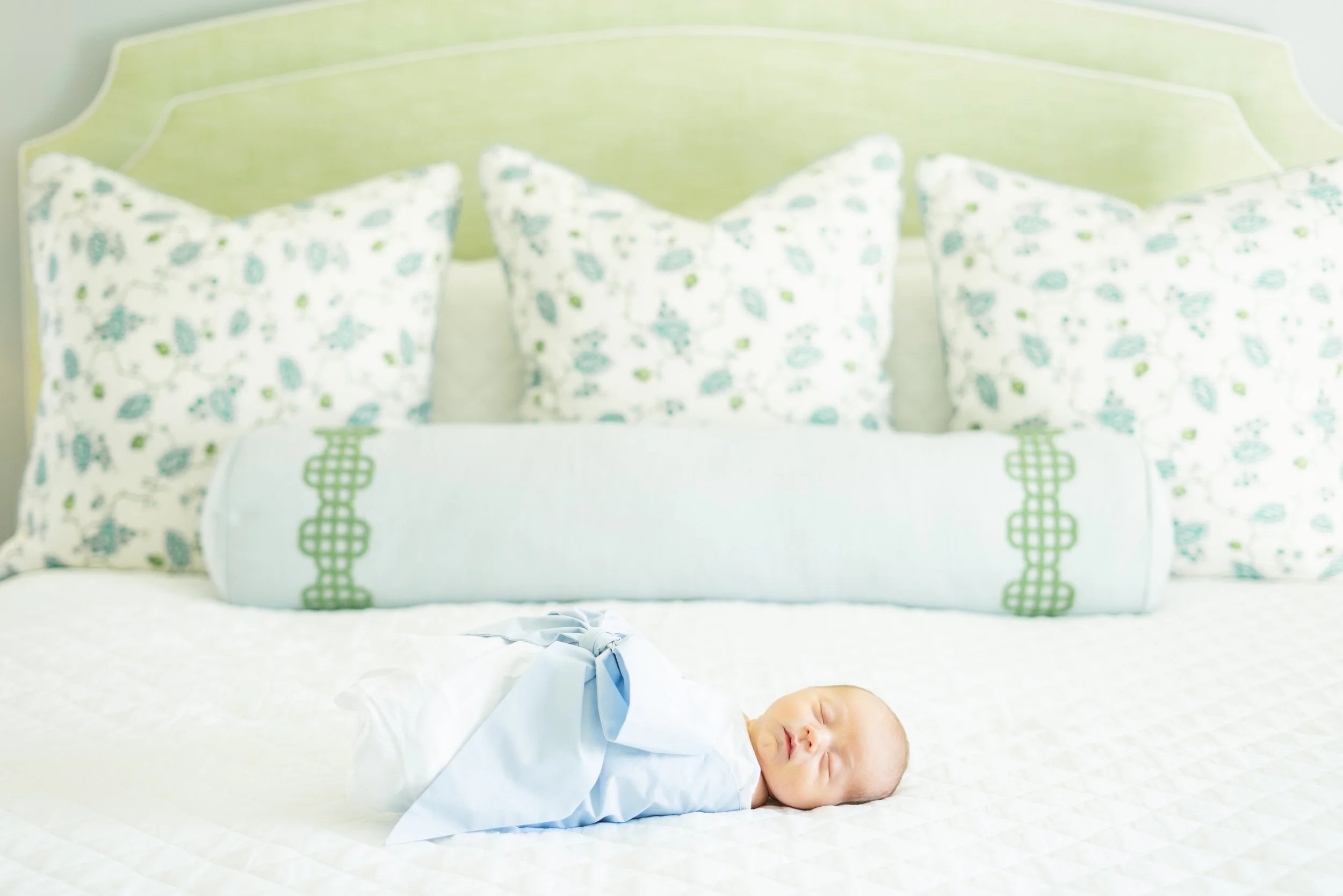 Sleeping baby in white pajamas lying on a bed with patterned pillows behind.