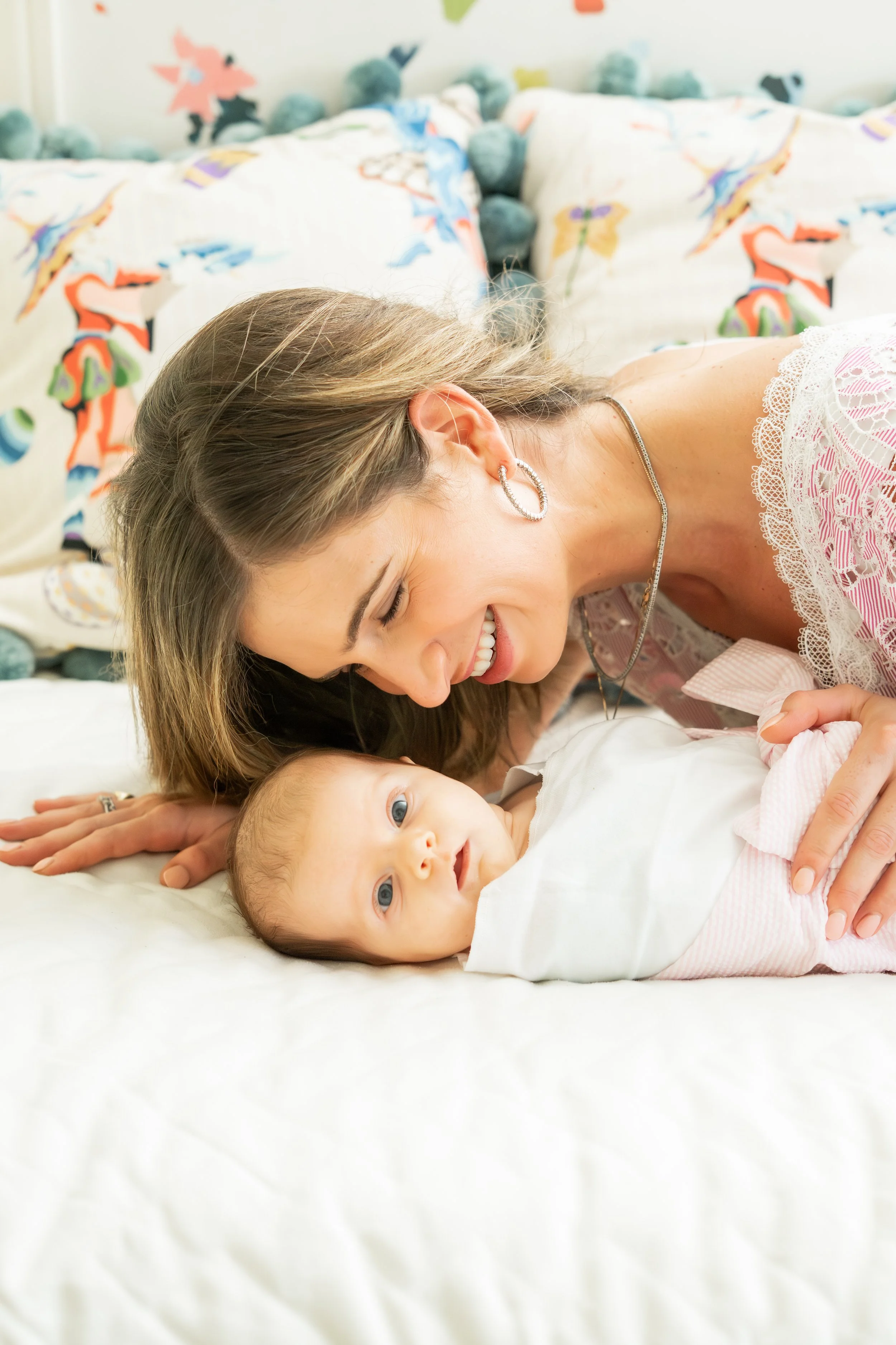 A woman with long brown hair smiling and leaning over a young boy with blond hair, who is lying on a white bed with his head turned and looking at the camera. The background features pillows with colorful, abstract animal designs.