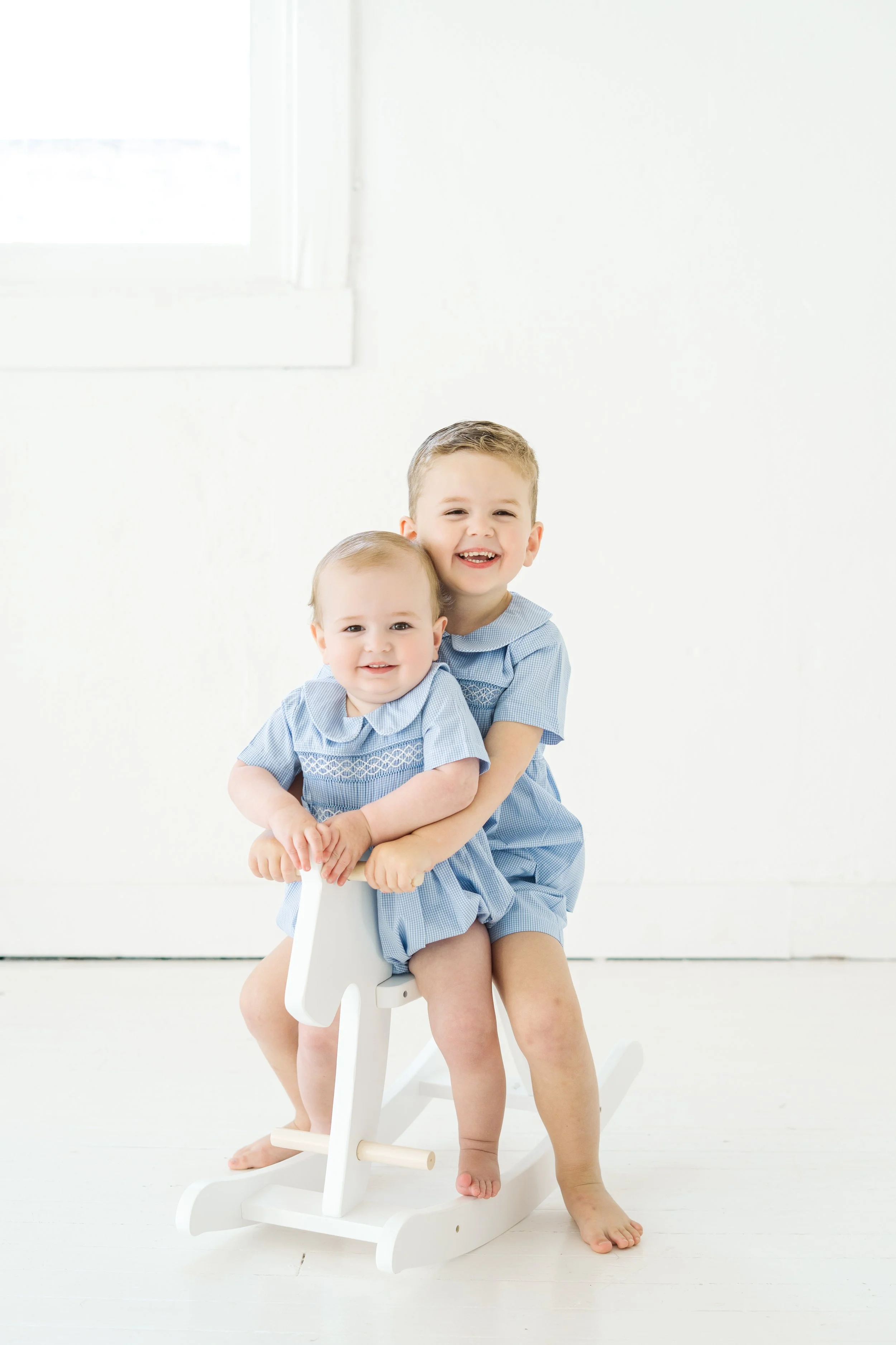 Two young boys with light hair and light skin wearing light blue clothing, sitting on a white rocking horse in a bright room with white walls and a window.