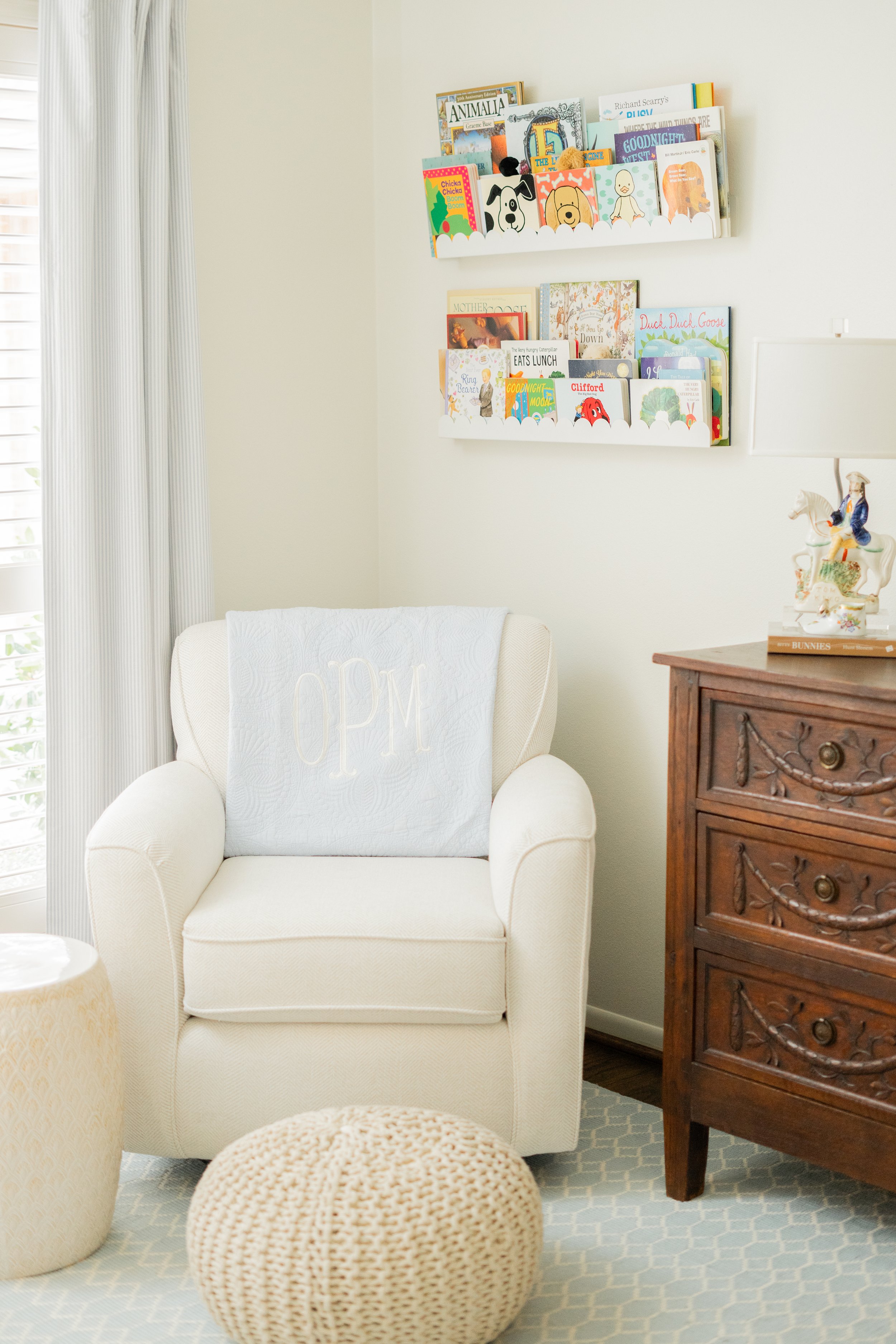 A cozy living room with a white armchair, a knitted pouf, a wooden dresser, and wall-mounted bookshelves filled with children's books. Light streams in through floor-to-ceiling curtains on the left.