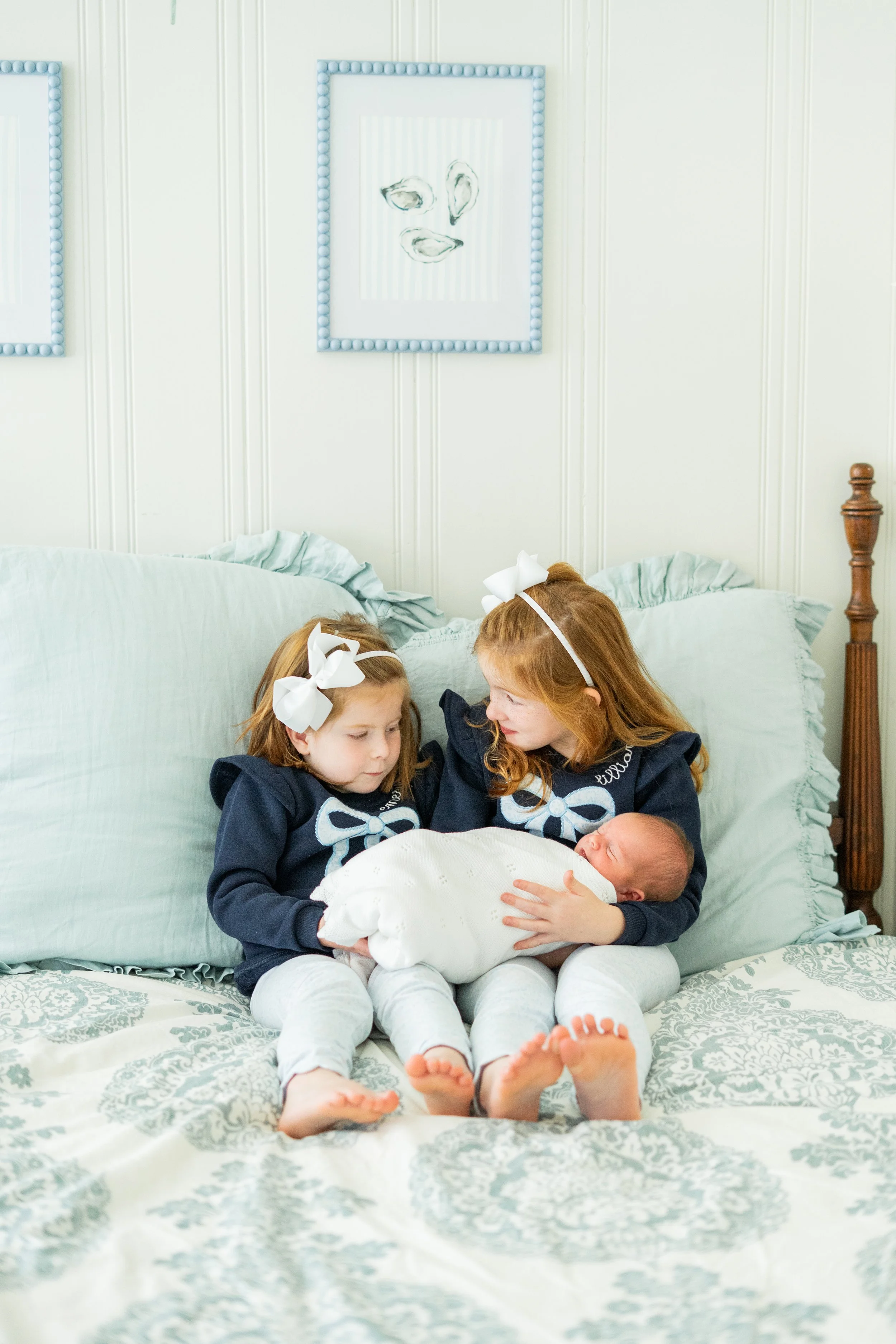 Two young girls with bows in their hair sitting on a bed, holding a newborn baby wrapped in a white blanket.