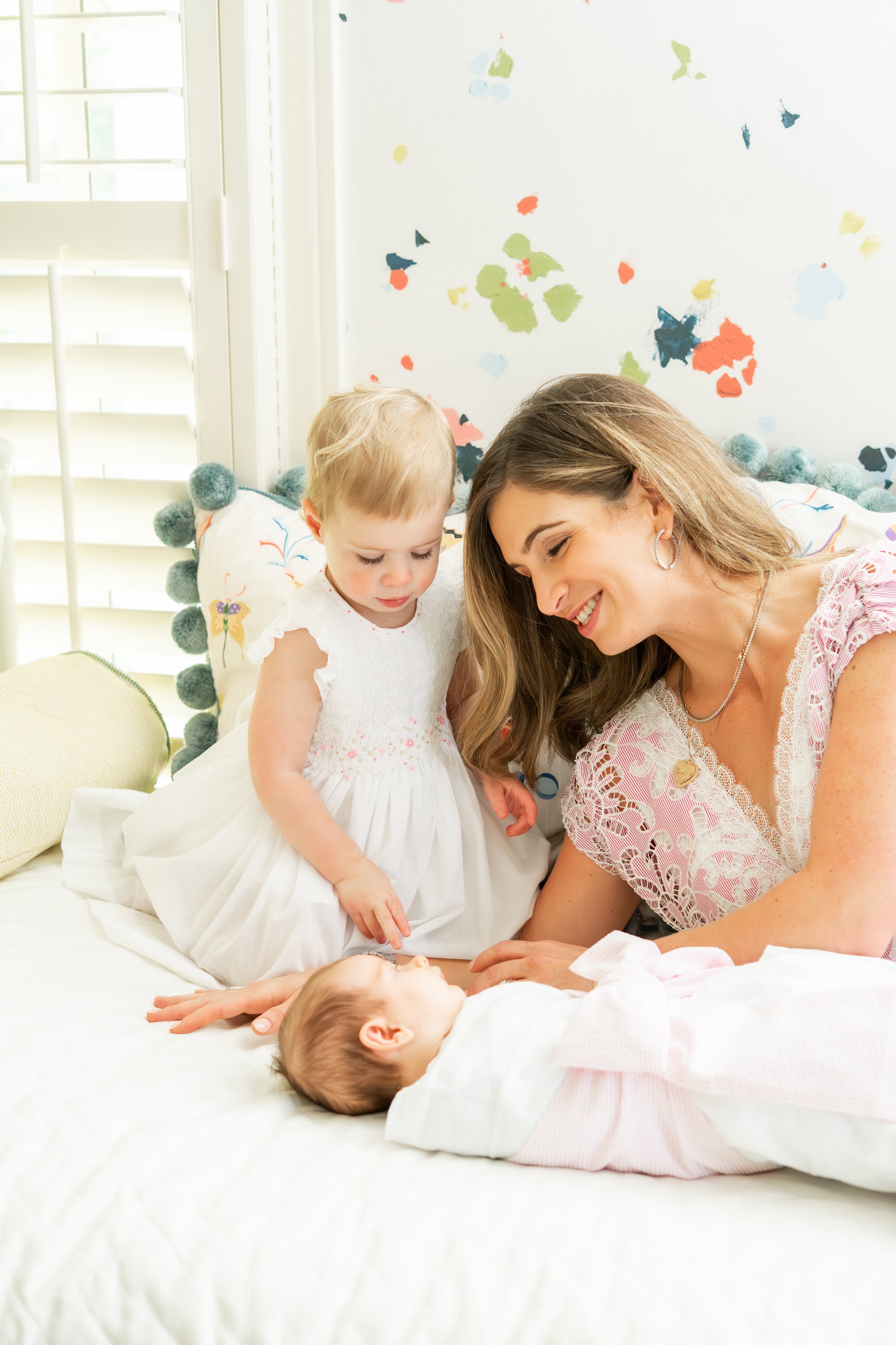 A woman and two young children, a girl and a boy, with light skin, are on a bed in a brightly lit room with white walls and blinds. The woman is smiling while watching the boy and girl, who are looking at an infant lying on the bed. The girl is weari
