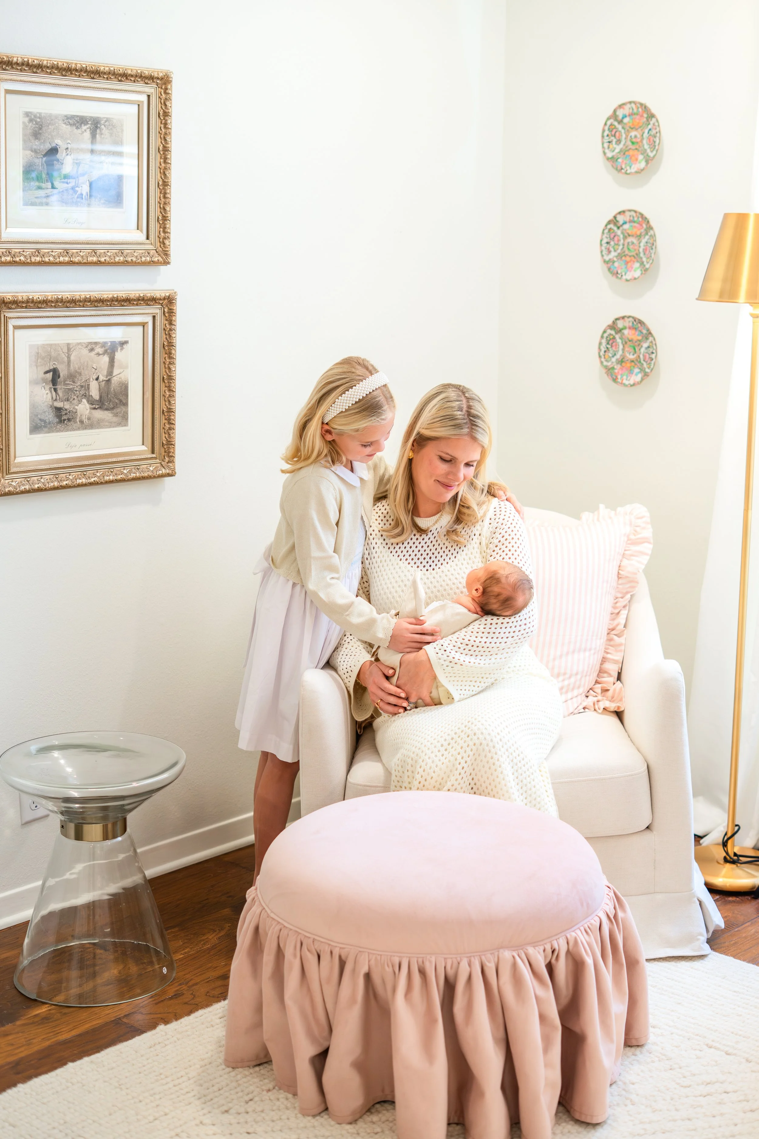 A woman holding a baby on her lap, a young girl standing next to her, all three sitting on a white armchair in a bright room. There are framed pictures and decorative plates on the wall.