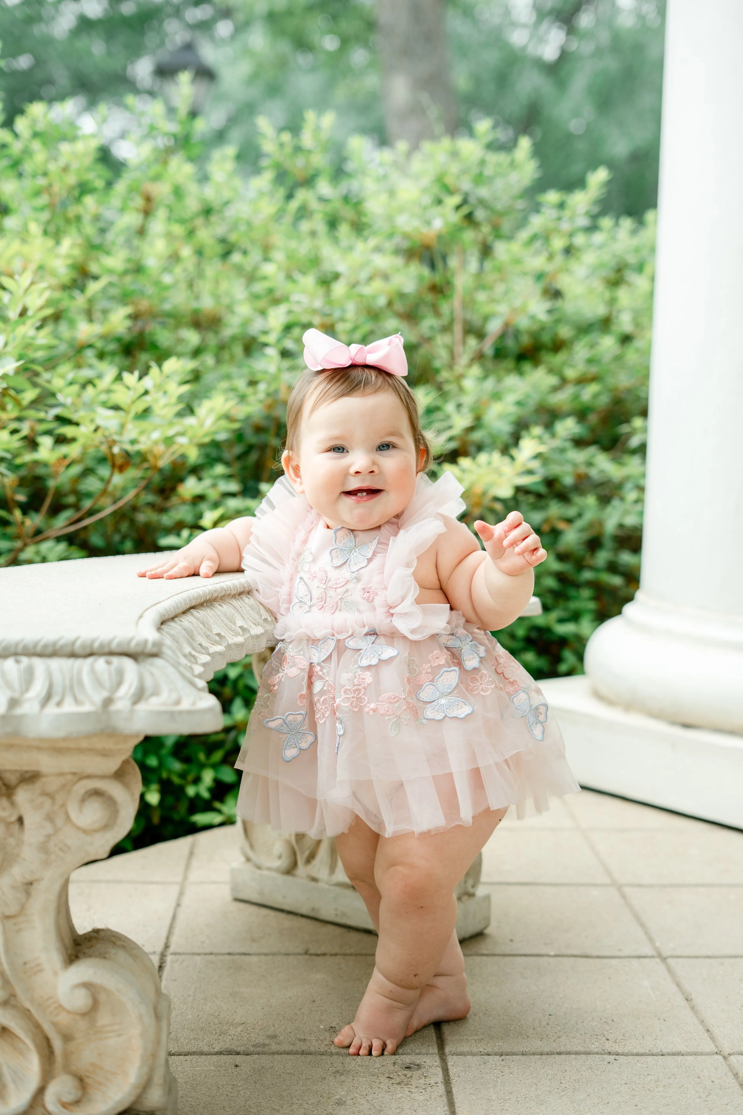 A smiling young girl wearing a pink dress with butterfly embroidery, a pink bow headband, and no shoes, standing outdoors on a tiled surface near a white ornate bench with greenery in the background.
