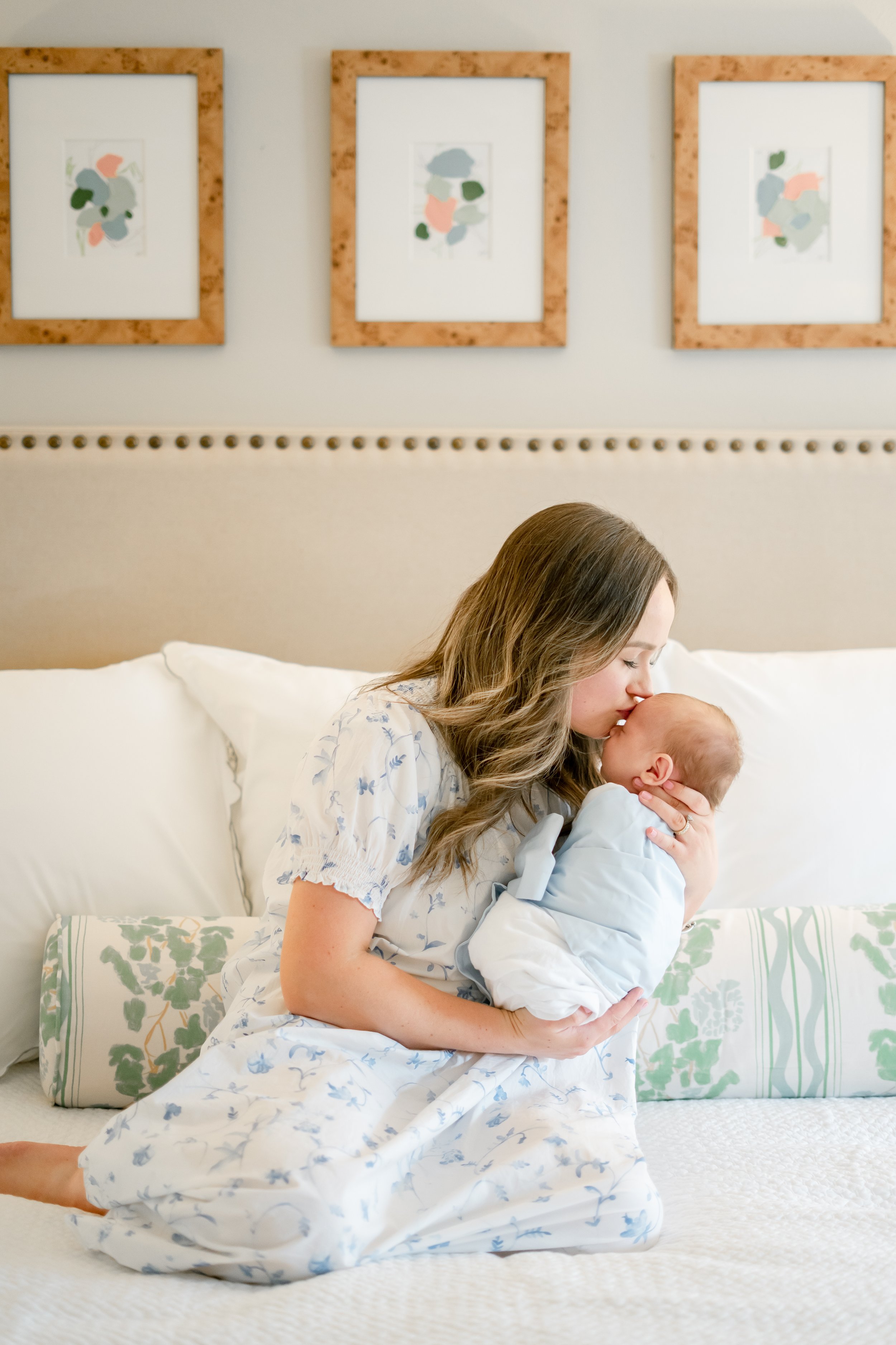A woman in a floral-patterned dress sits on a bed, holding a newborn baby, gently touching her face to the baby's forehead in a cozy bedroom with three framed abstract artwork pieces on the wall behind her.