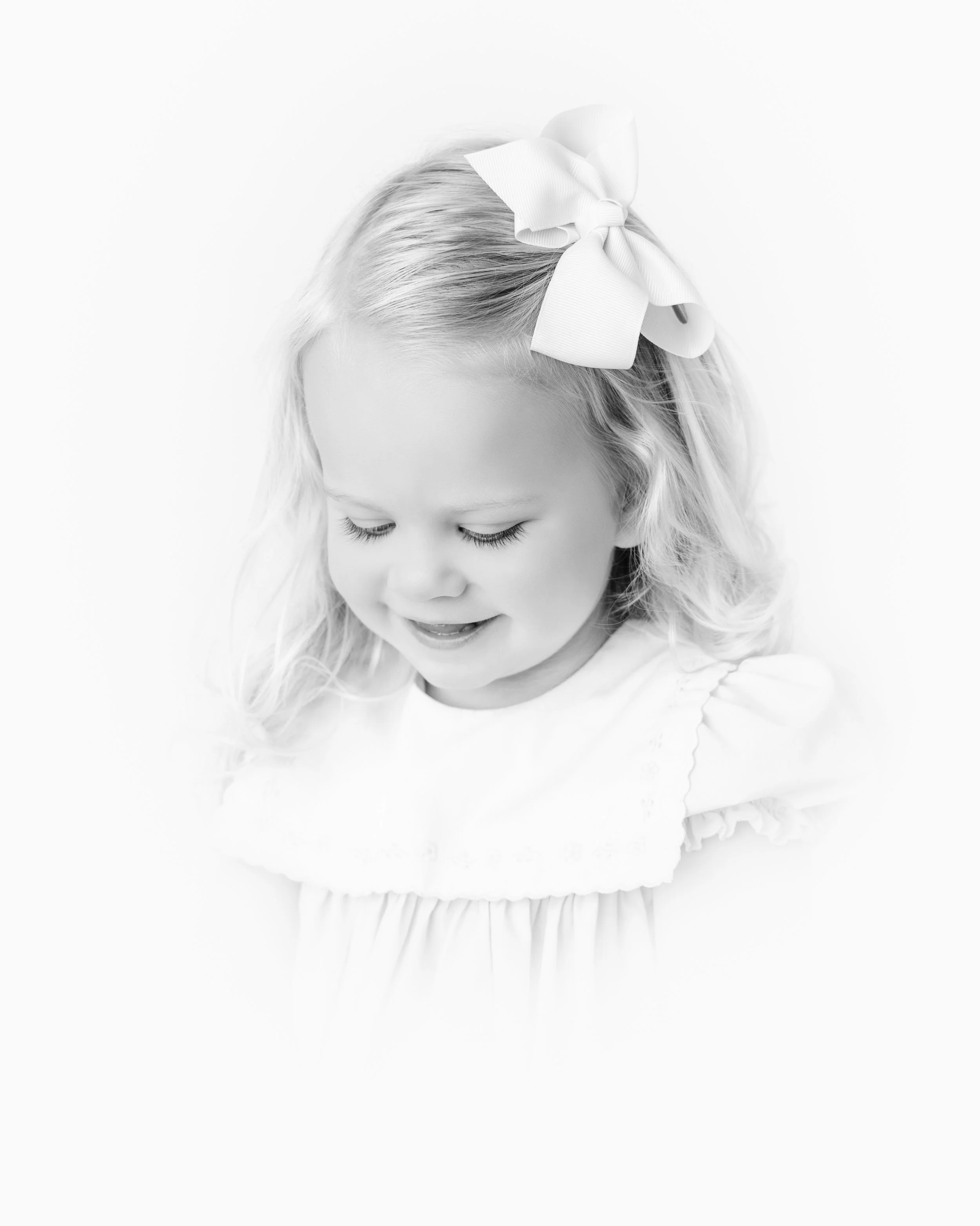 A young girl with light-colored hair and closed eyes, wearing a large white bow in her hair, dressed in a white top with ruffled sleeves, looking downward on a plain white background.