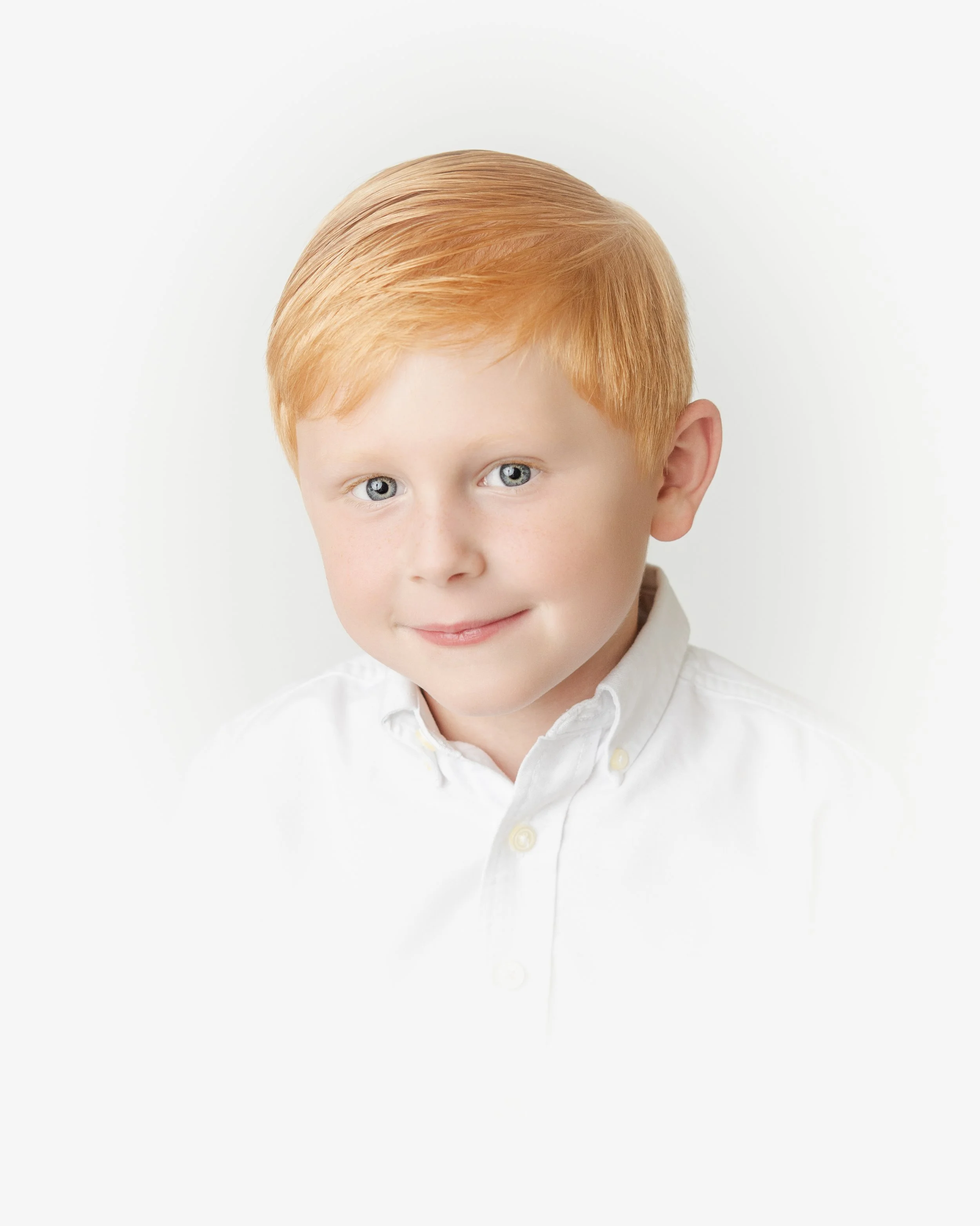 Close-up of a young boy with pigmented red hair, blue eyes, and a fair complexion, wearing a white shirt with a collar, against a plain white background.