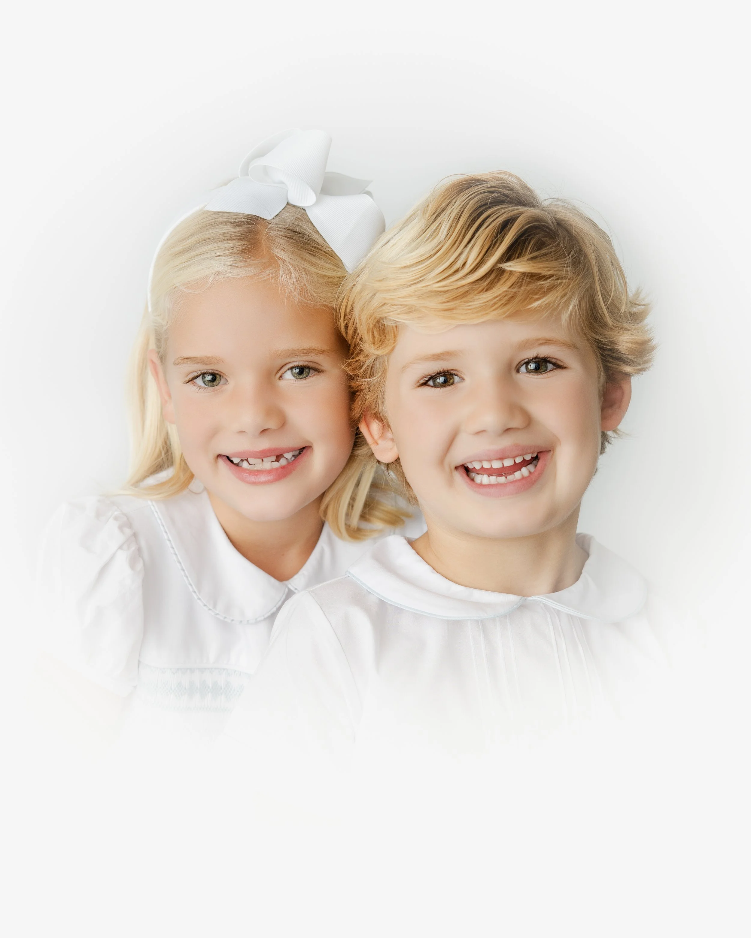Two young children, a blonde girl with a white bow on her head and a boy with wavy blonde hair, both smiling and wearing white tops, close together against a plain white background.