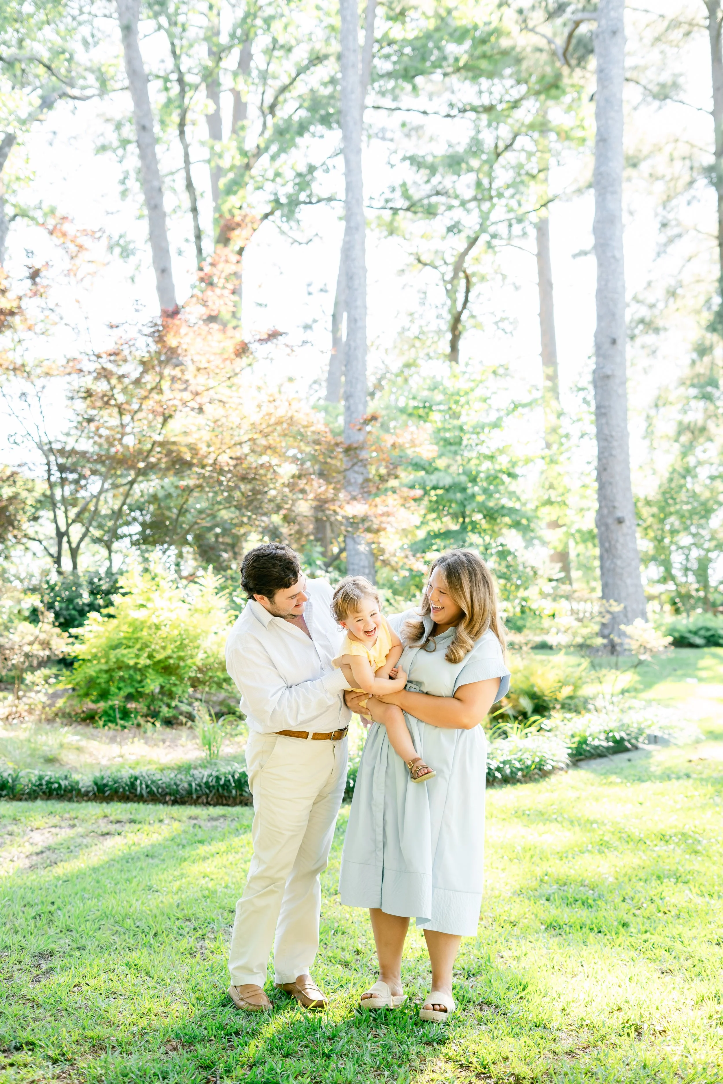 A family of three, a man, a woman, and a small child, standing in a lush green garden with trees and bushes in the background. The woman is holding the child, and they are all smiling and looking at each other on a sunny day.