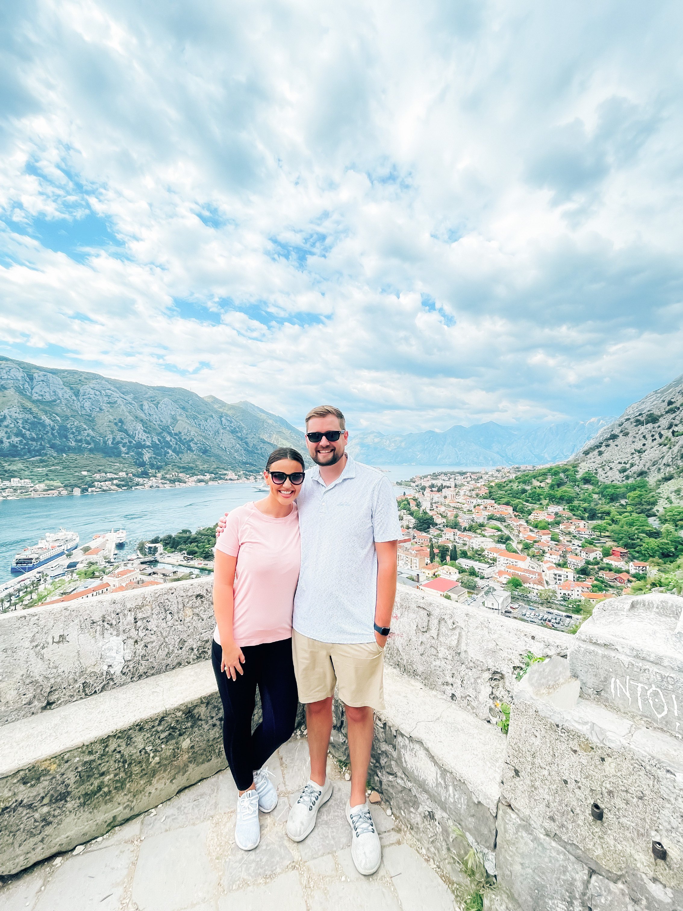 A smiling couple stands on a stone ledge overlooking a waterfront city with mountains in the background. The woman wears sunglasses, a light pink shirt, and black pants. The man wears sunglasses, a white patterned shirt, and khaki shorts.