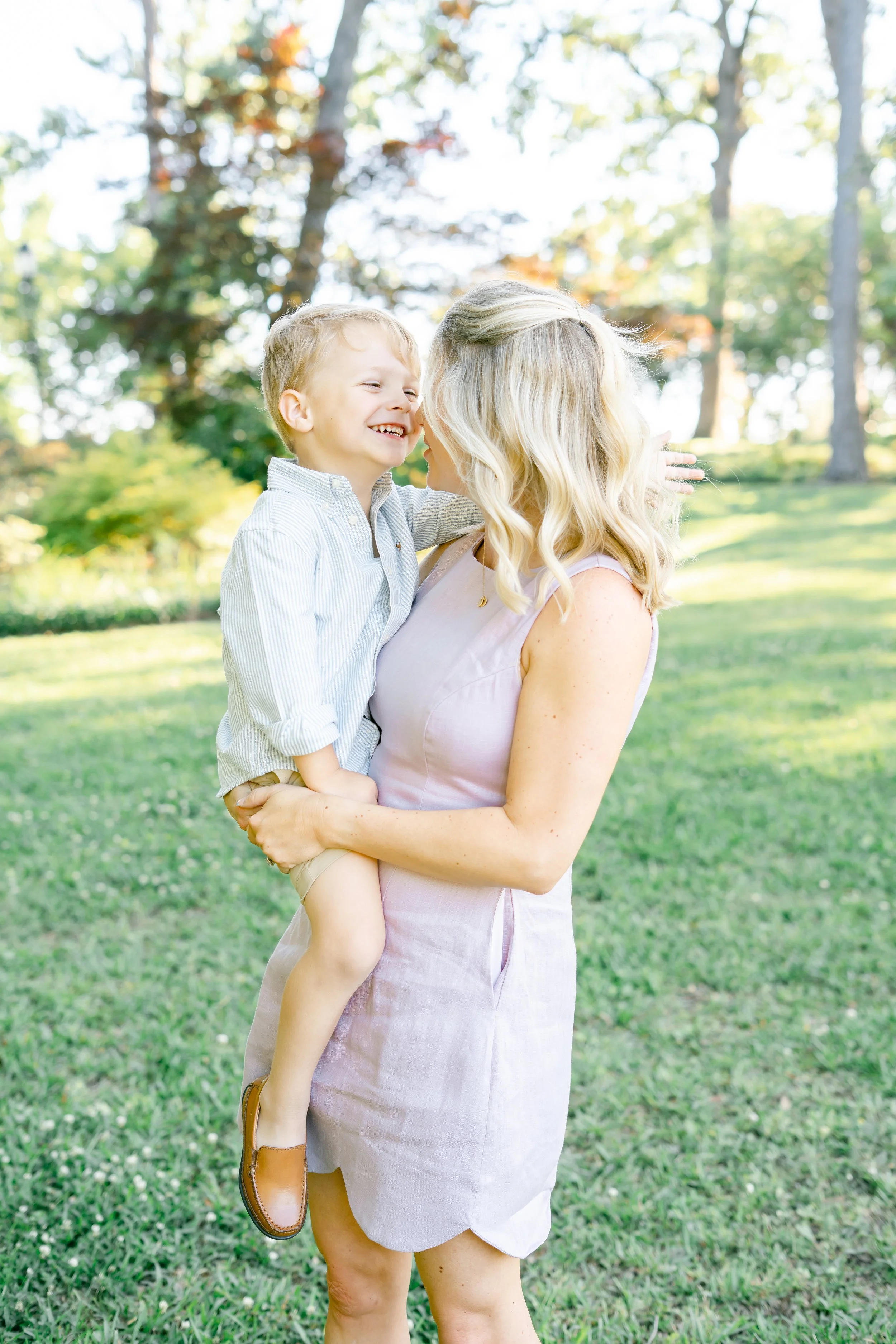 A woman with blonde curly hair wearing a light pink dress holding a young boy with blonde hair in a light striped shirt and beige shorts outdoors in a park with trees and grass.