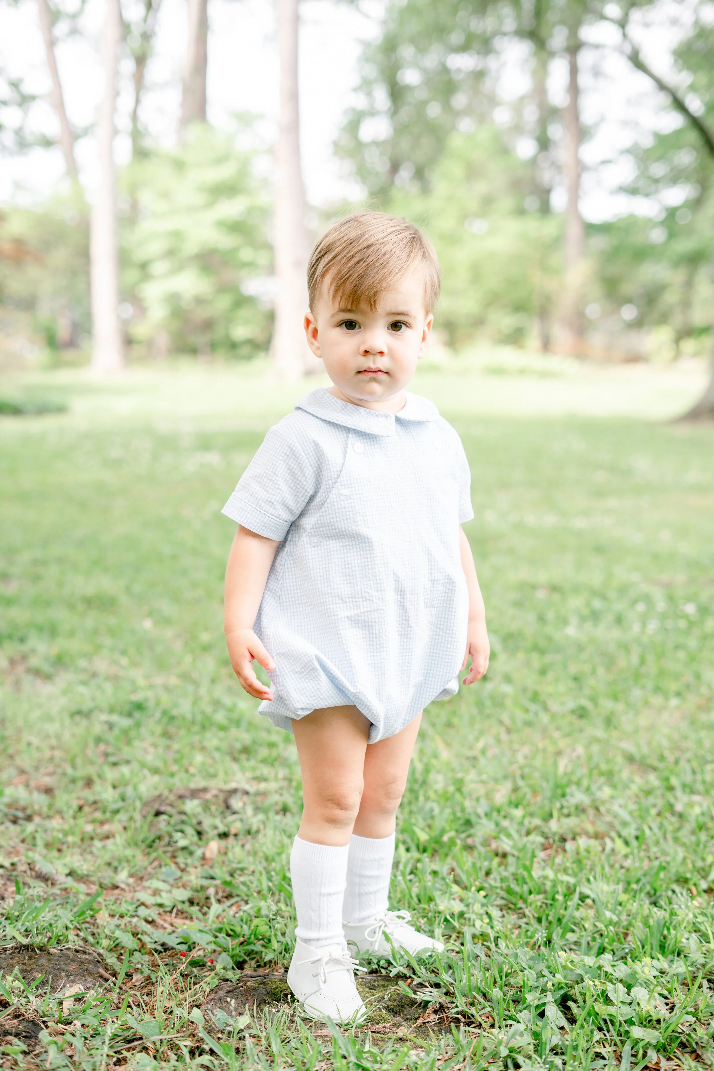A young boy with short light brown hair and fair skin standing outdoors on grass, wearing a light blue jump-suit, white knee-high socks, and white sneakers, with a background of trees and greenery.