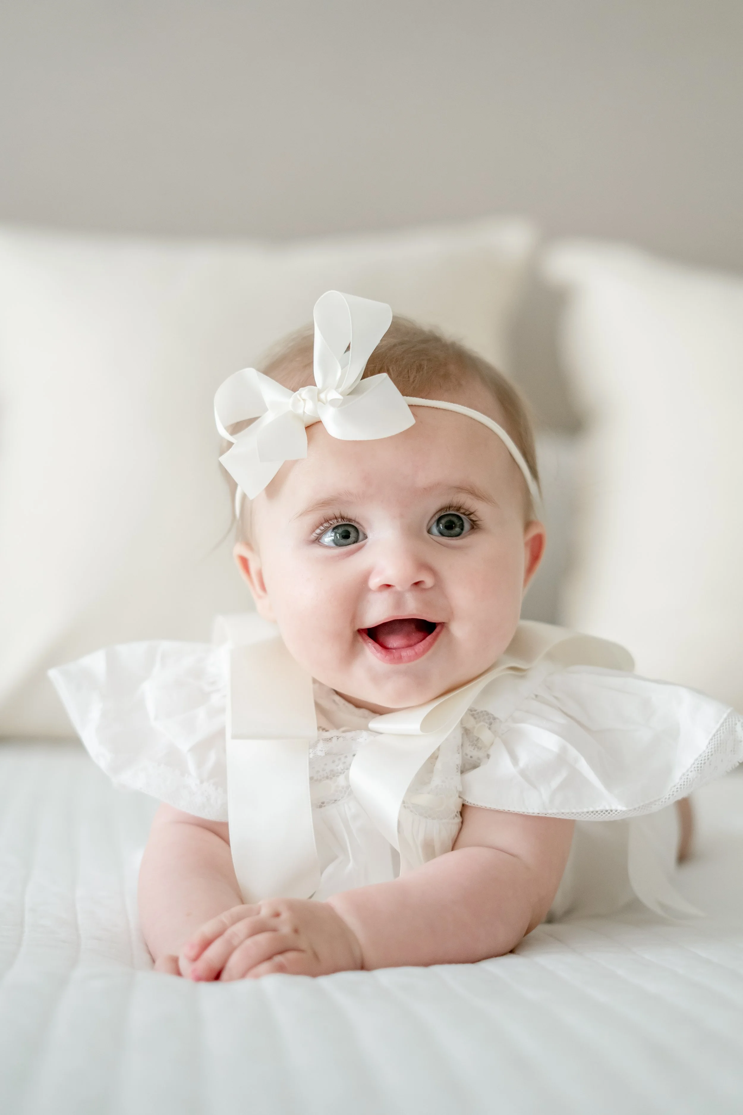 Young girl with blue eyes and blonde hair, wearing a white dress and a white headband with a bow, smiling and lying on a white bed.
