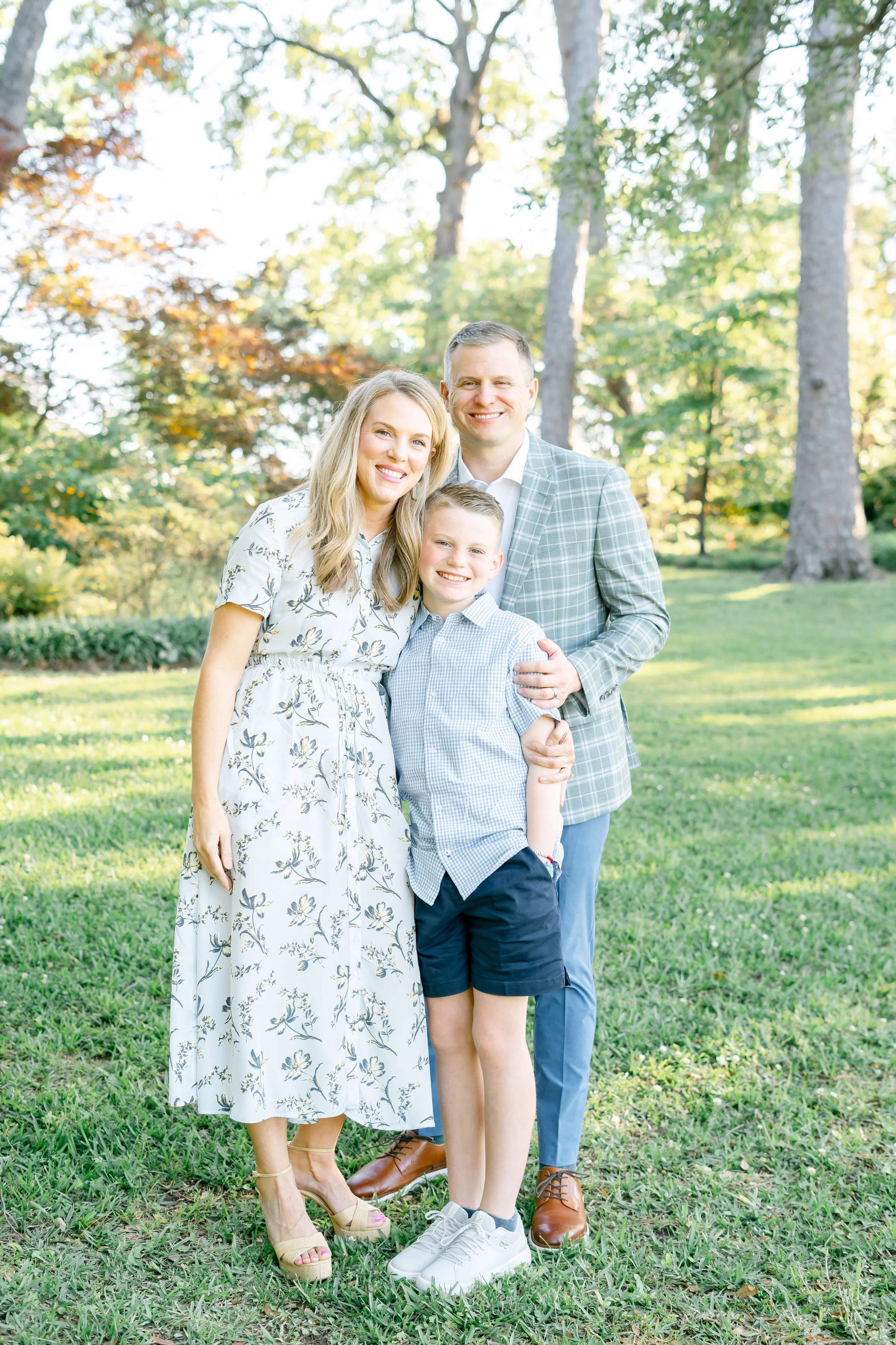 A family of four standing on grass in a wooded park with trees and sunlight in the background. The mother wears a white floral dress and beige high heels. The father wears a gray plaid blazer, white shirt, and brown dress shoes. The son wears a light