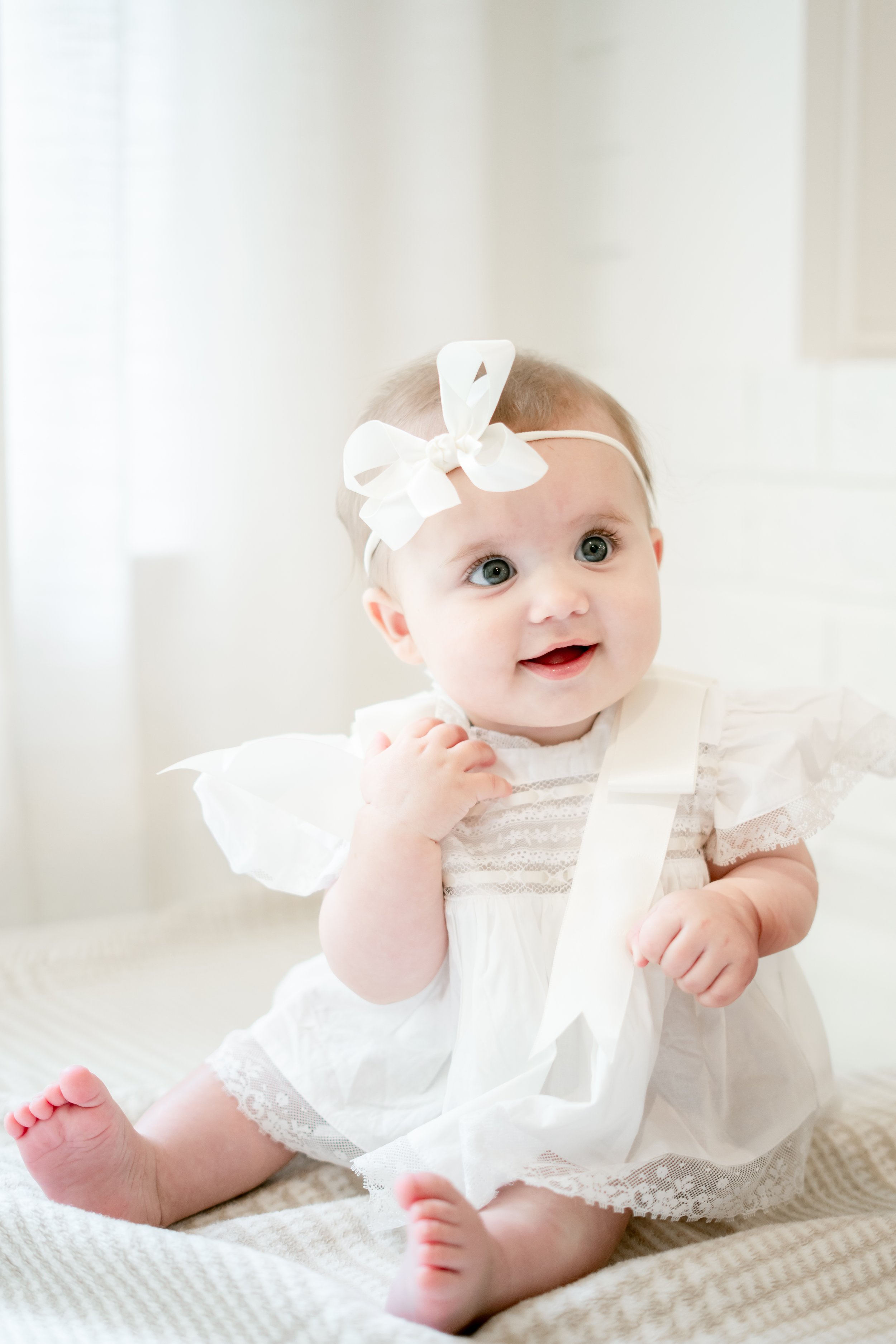 A baby girl with blue eyes and blonde hair, wearing a white dress with lace details and a large white bow headband, sitting on a beige and white striped blanket.