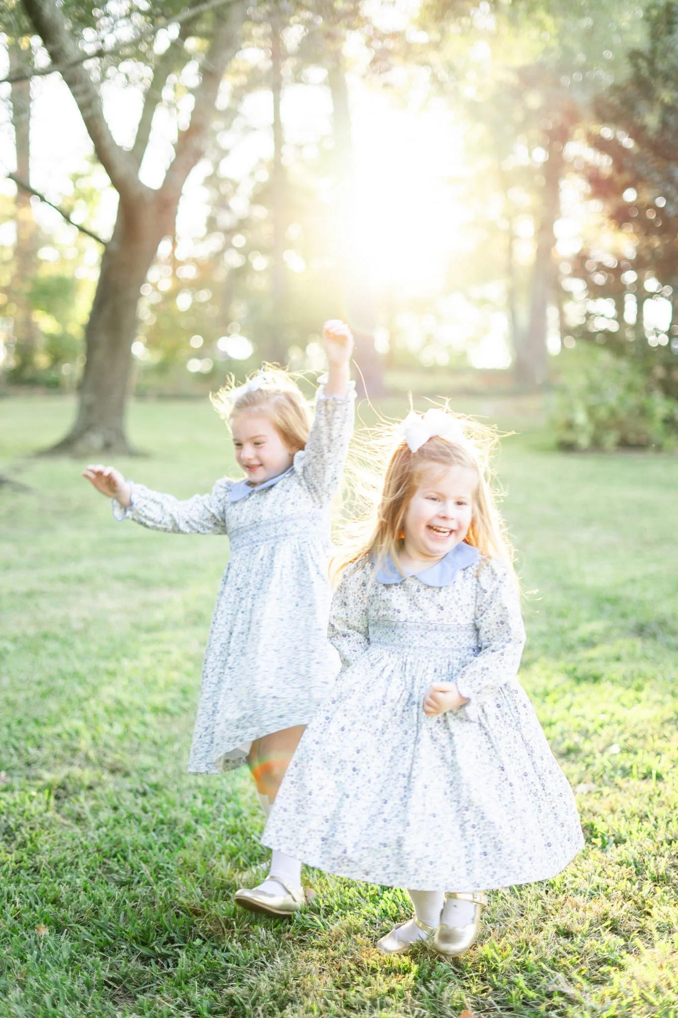 Two young girls in floral dresses playing outdoors on a grassy field with trees in the background, illuminated by bright sunlight.