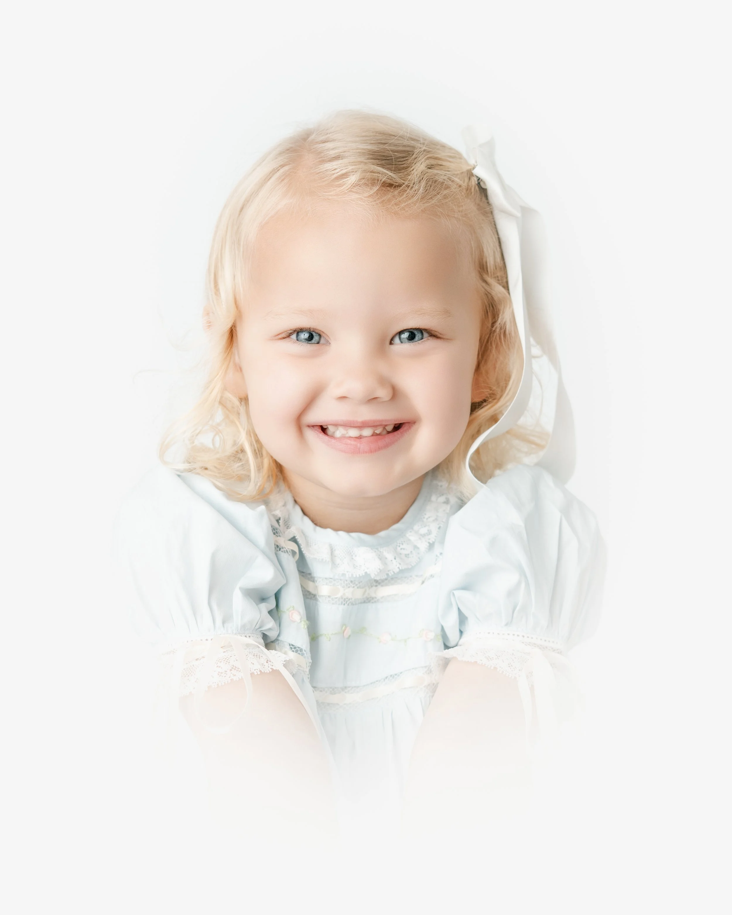 Close-up of a smiling young girl with blonde hair and blue eyes, wearing a white dress with lace details, sitting against a white background.