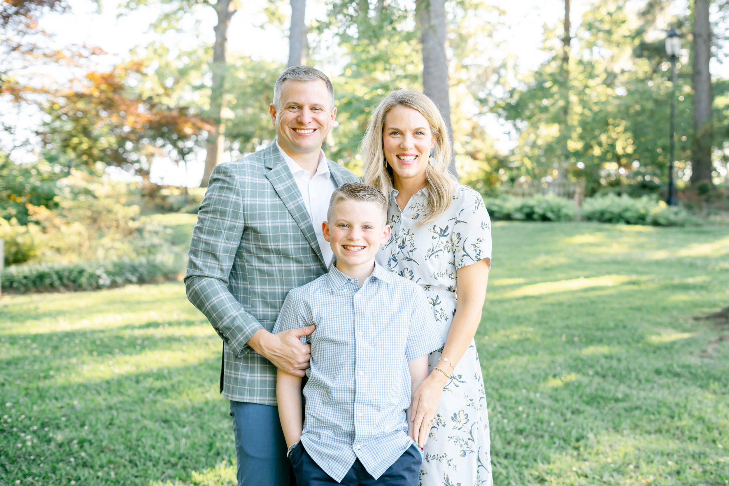 A smiling family of four posing outdoors in a park with green trees in the background. The family includes a man, woman, and two boys, all dressed in light-colored and checkered clothing.