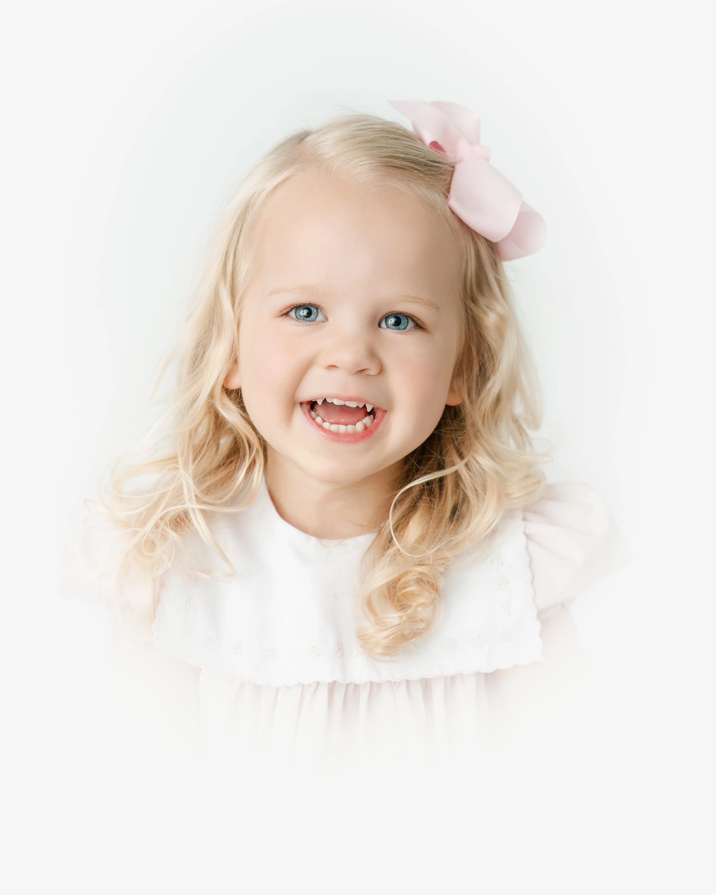 Close-up portrait of a young girl with blonde hair, blue eyes, and a wide smile showing missing front teeth, wearing a pink bow in her hair, against a white background.
