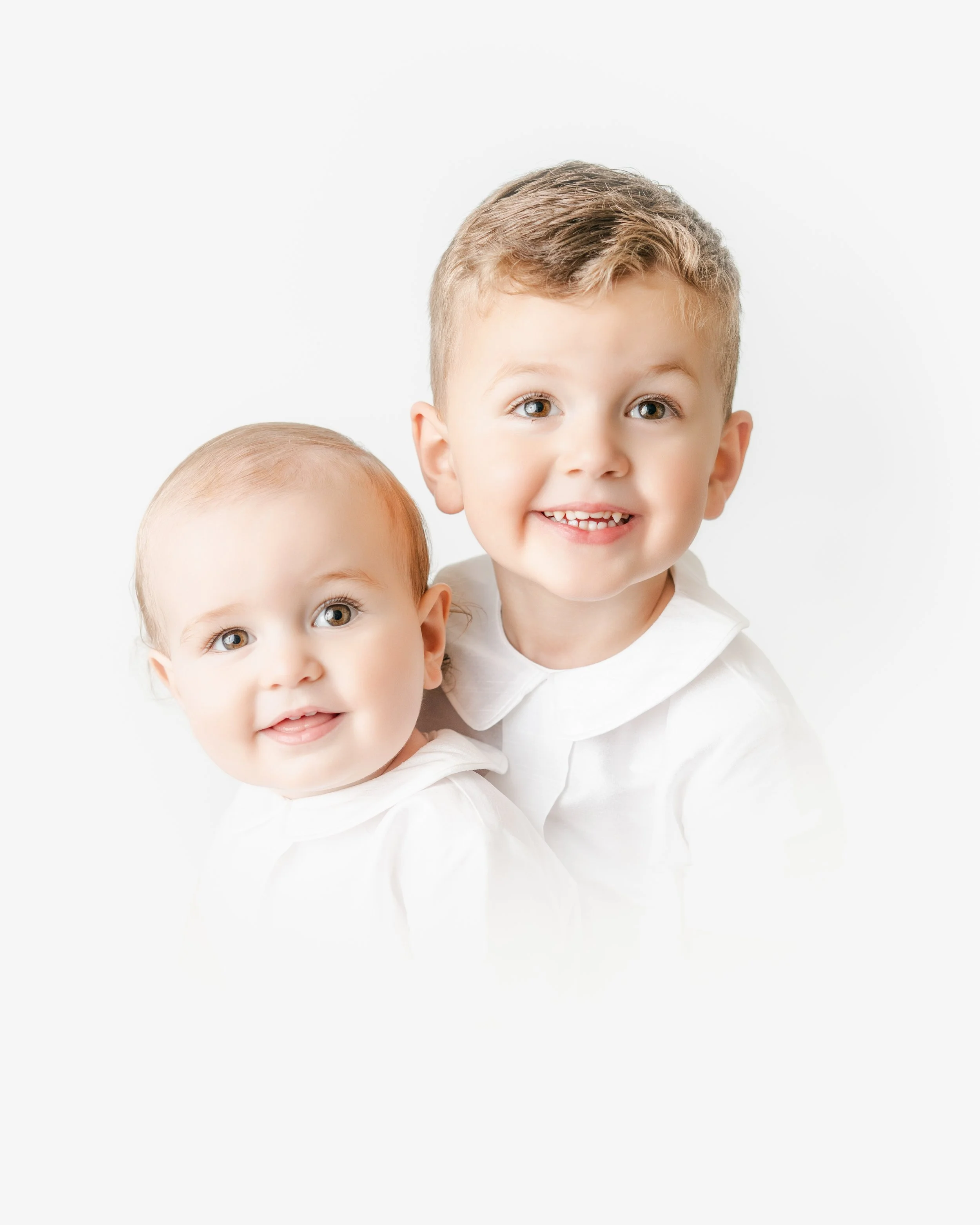 Close-up of two smiling young children with curly blonde hair and blue eyes, wearing white shirts against a plain white background.