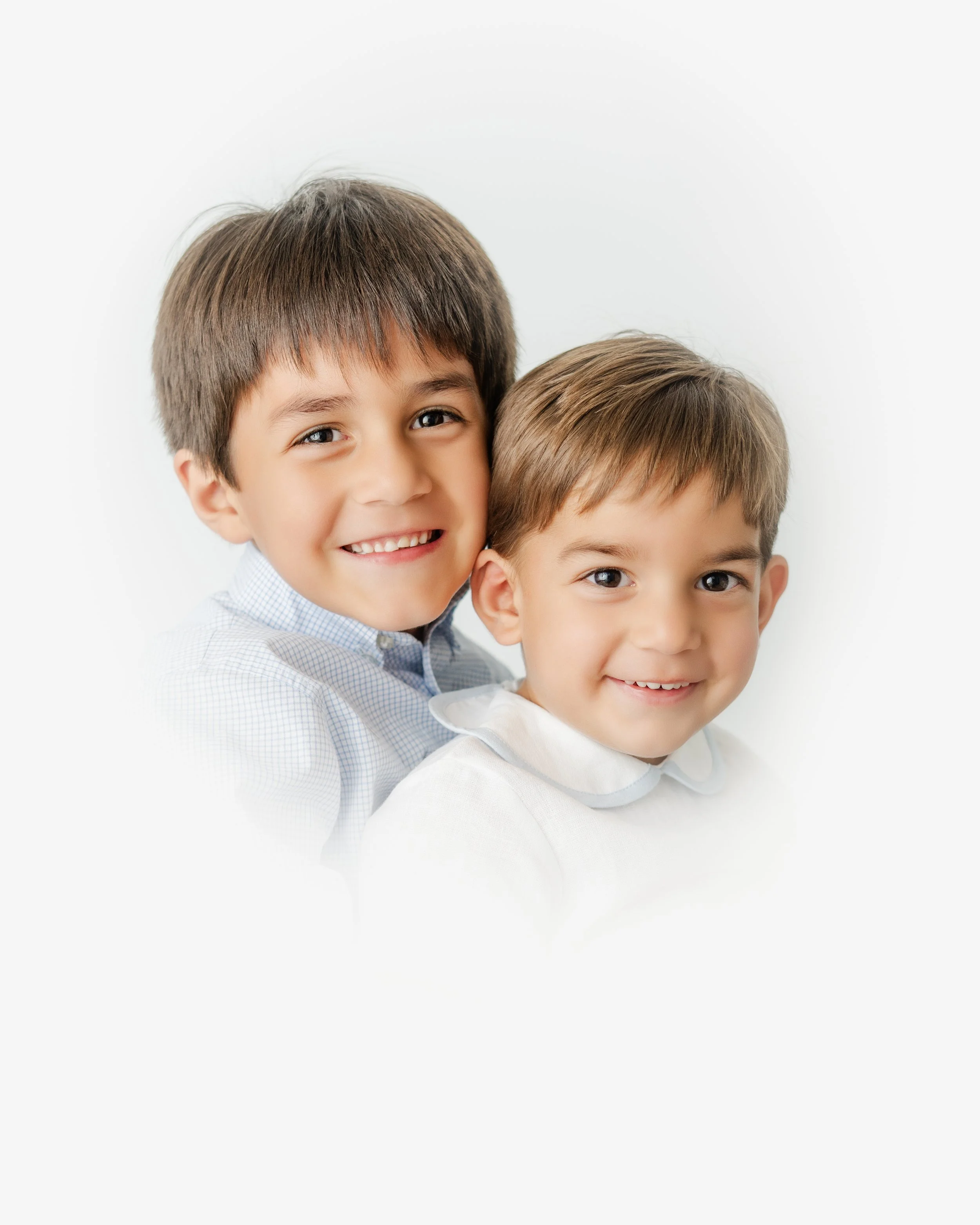 Two young boys with brown hair smiling and looking at the camera, wearing light-colored shirts, against a plain white background.