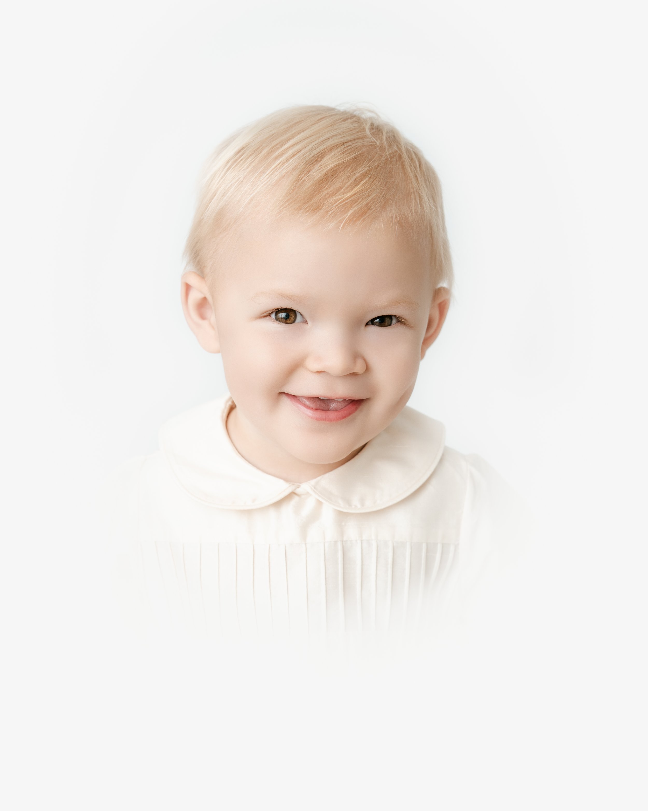 Close-up of a smiling young boy with light blonde hair and fair skin, wearing a cream-colored top against a plain white background.