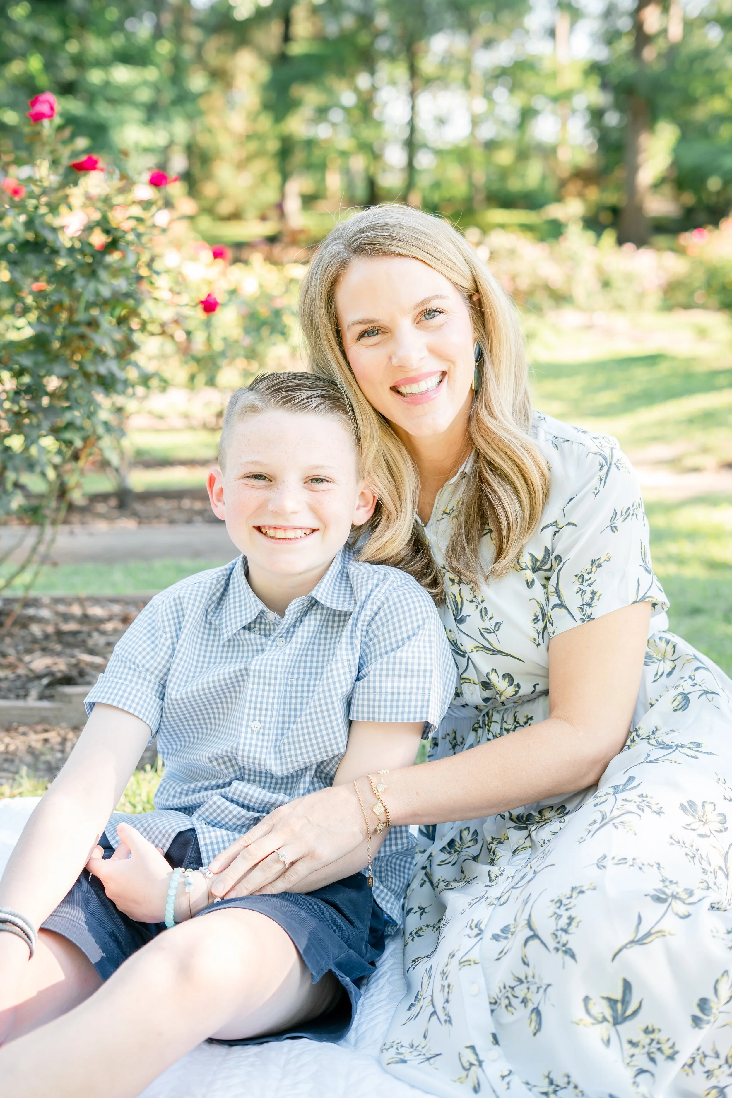 A woman and a boy sitting outdoors in a garden with blooming roses and trees, smiling at the camera on a sunny day.